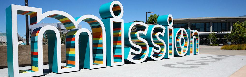 Mission College sign on campus. Large white letters with the outside painted turtoise and the interior painted orange, yellow, red, teal, and light green in alternating stripes.