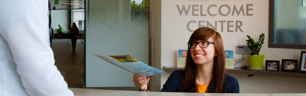 Young woman with straight brown hair and black-framed glasses hands a brochure to a potential student from behind her desk. Behind her, a large sign that reads "Welcome Center" is visible. 