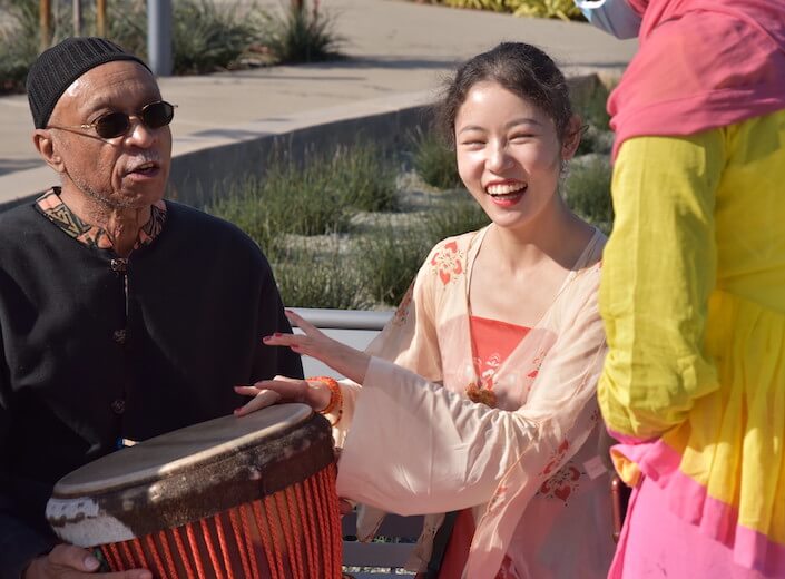 International Day - a young woman of Asian descent in traditional Southeast Asian clothing drums on a Djembe belonging to Dr. Piper (an older man of African-American descene in traditional clothing.)