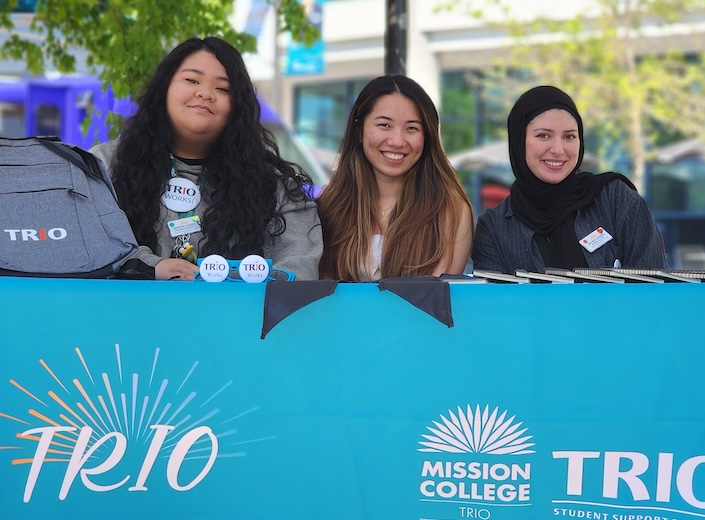 Three young women of color are sitting at TRIO booth on sunny day. One wears a jihab, the others have olive skin and long dark hair.