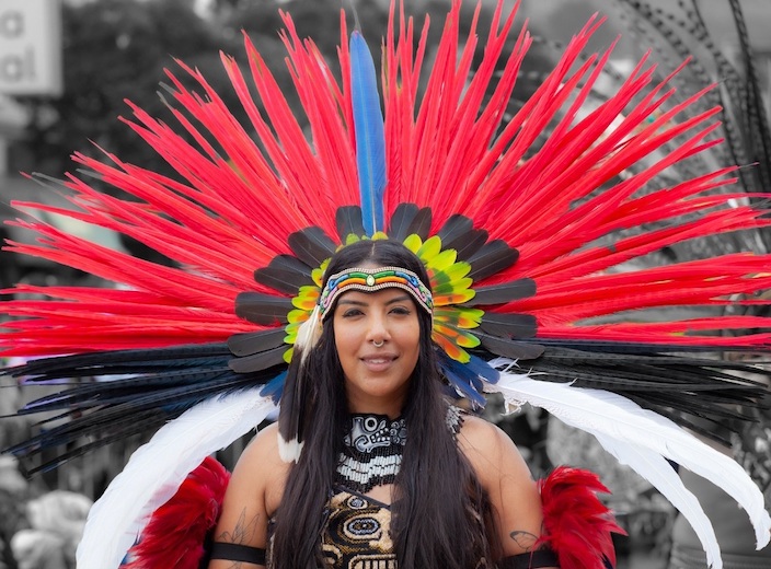 Sandra Quintero, a young Mexican-American owman with straight black hair wears a traditional Aztec dance costume with large red and b,ac feathers.