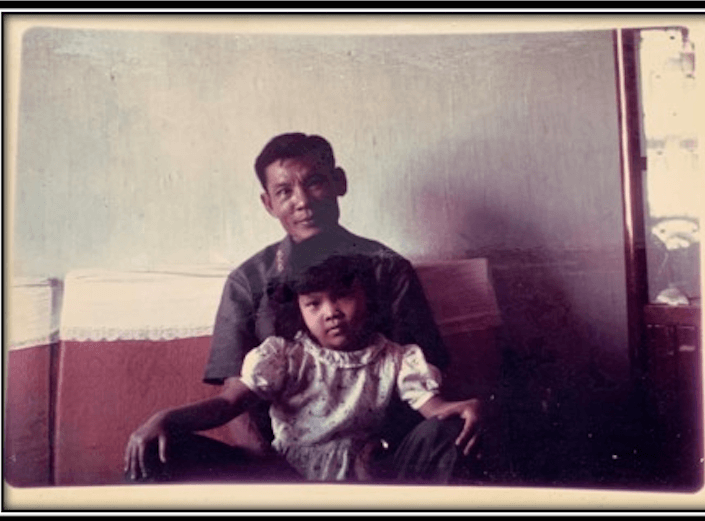 An aged photo from the seventies depicts a father and young female child on a couch. The girl raises her hands above her head. They are Vietnamese.