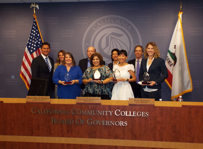 Julie Vu poses with an award with a group of colleagues in front of the California Community College seal in a formal meeting room.