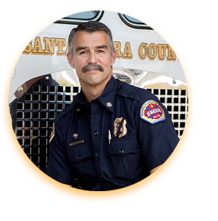 Ken Kehmna, a retired Fire Chief, poses in uniform against the front of a fire truck. He has a dark moustache and short greying hair. He is lean and fit.