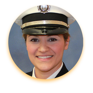 Soulmaz Brazil, a young woman posing in a fire service uniform and smiling. Her dark hair is pulled up under her military-style hat, and her skin is tan.