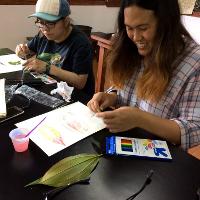 Two female students work with colored pencils, paper, and leaves in a classroom.