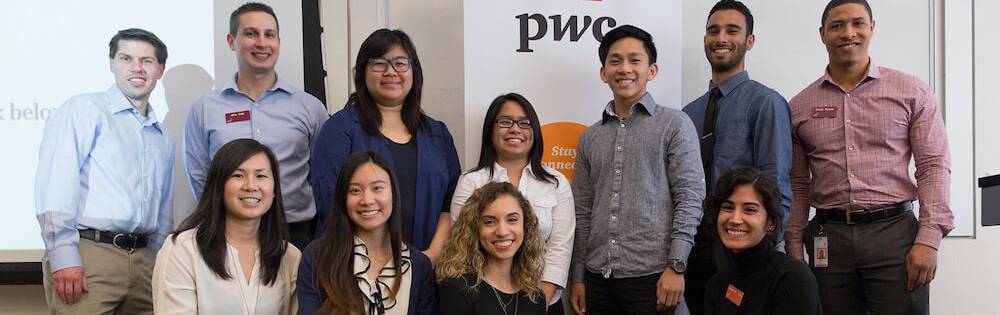 A group of college students stand together in fron of a sign that says PWC Accounting. They were business casual clothes.