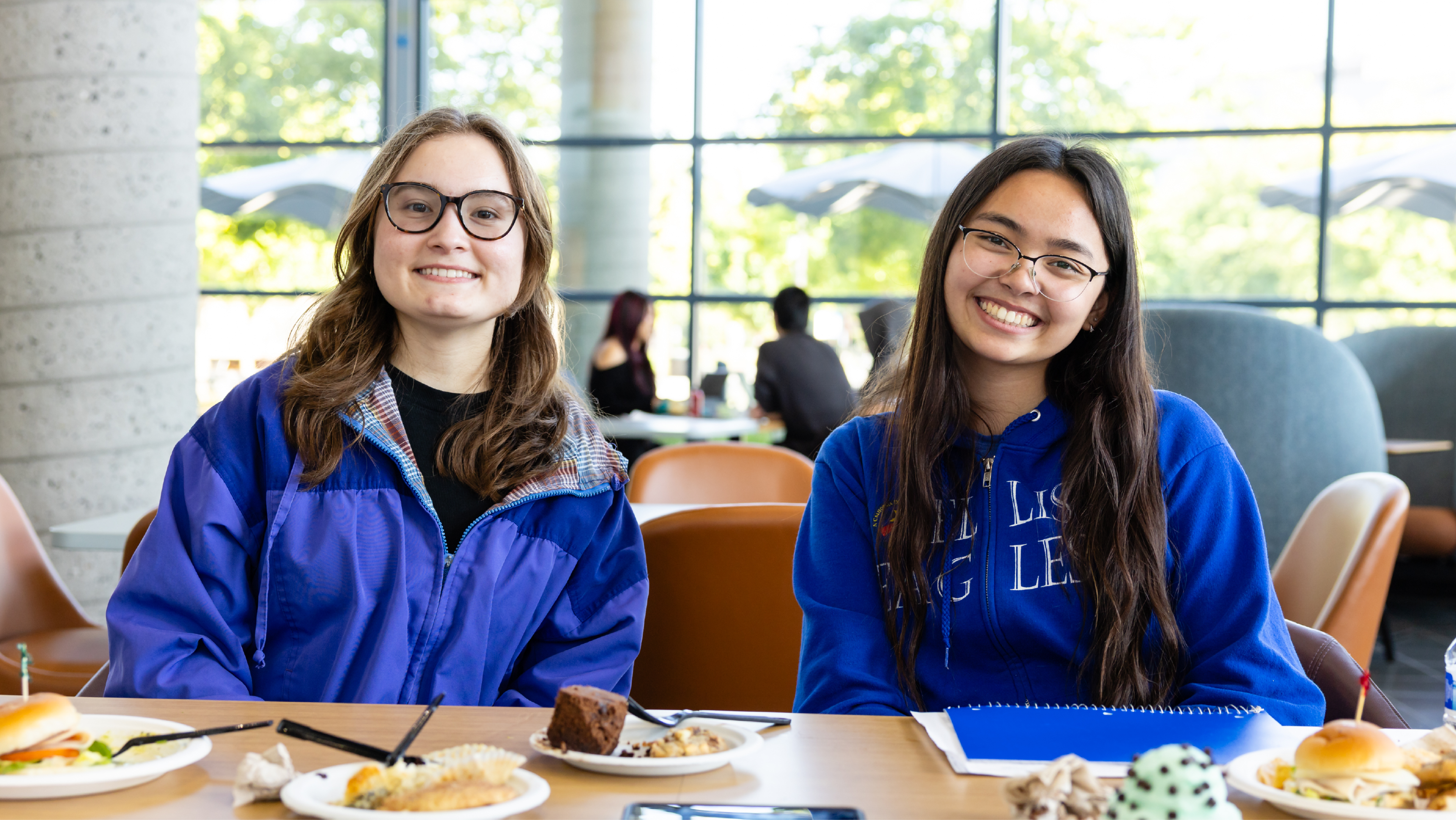 Two female students smiling at a dining table with plates of food and drinks in front of them.