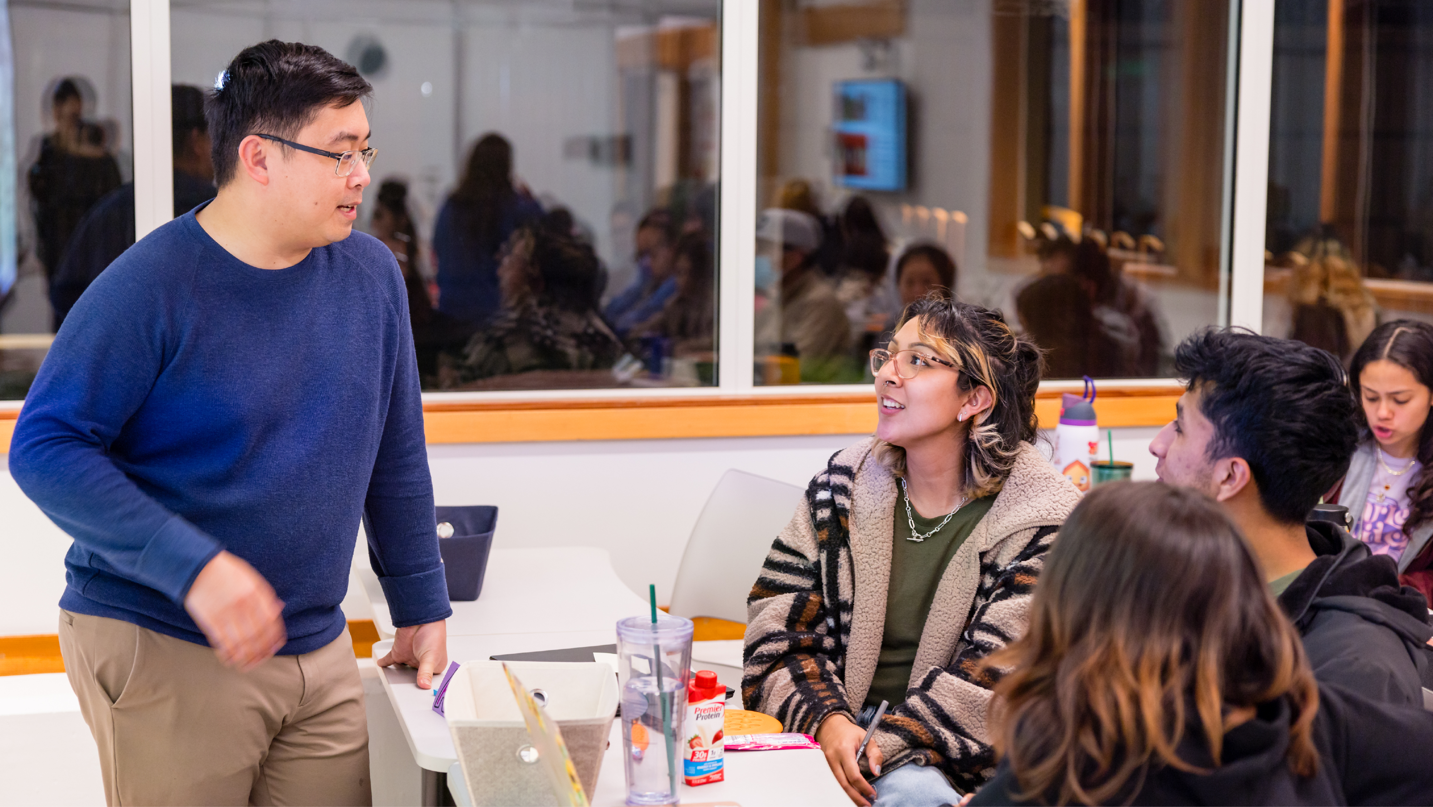 A male instructor standing and interacting with a small group of students seated at a classroom table.