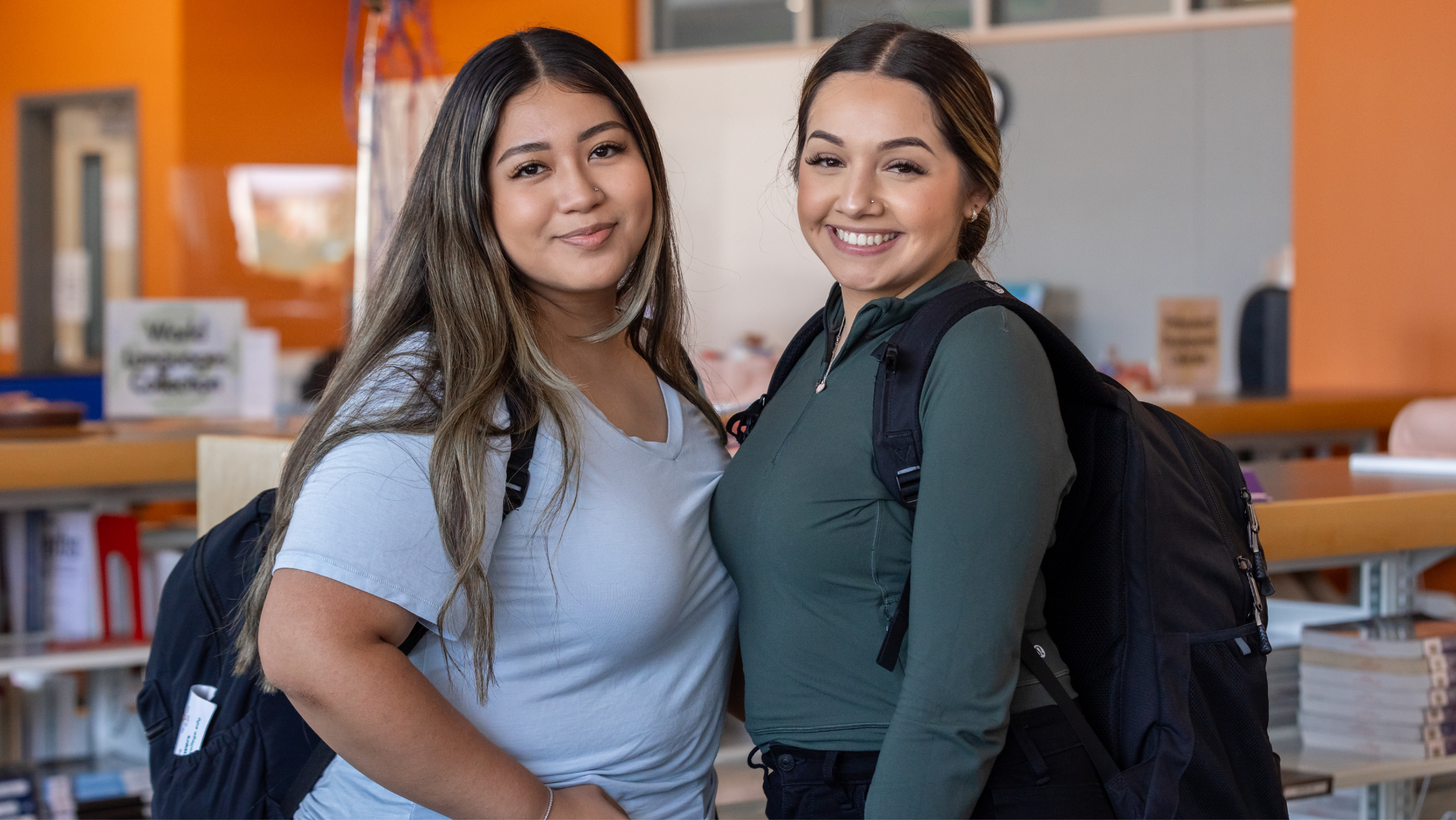 Two female students wearing backpacks smiling together indoors.