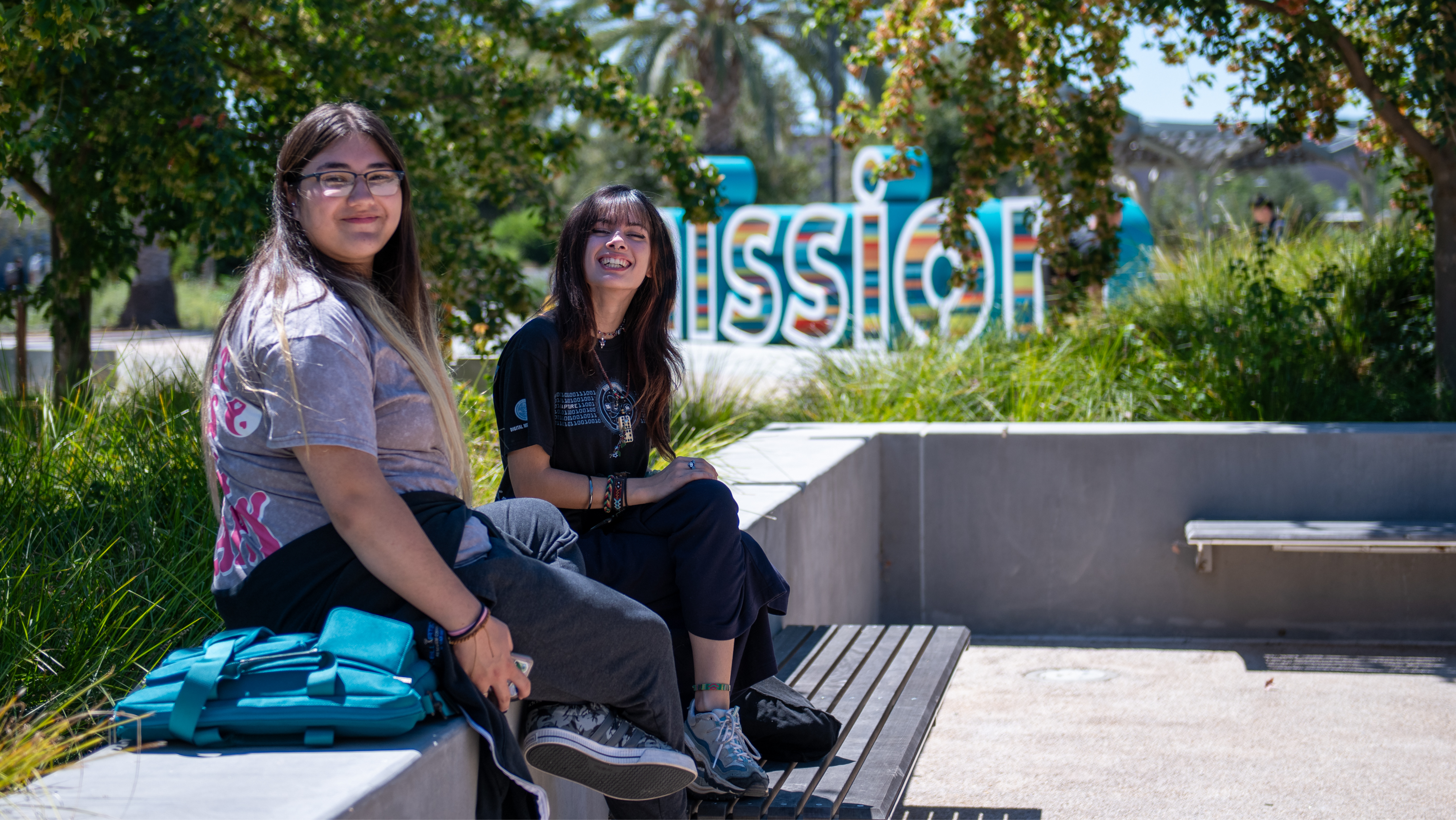 Two students sitting on an outdoor stone bench with the Mission College sign in the background.