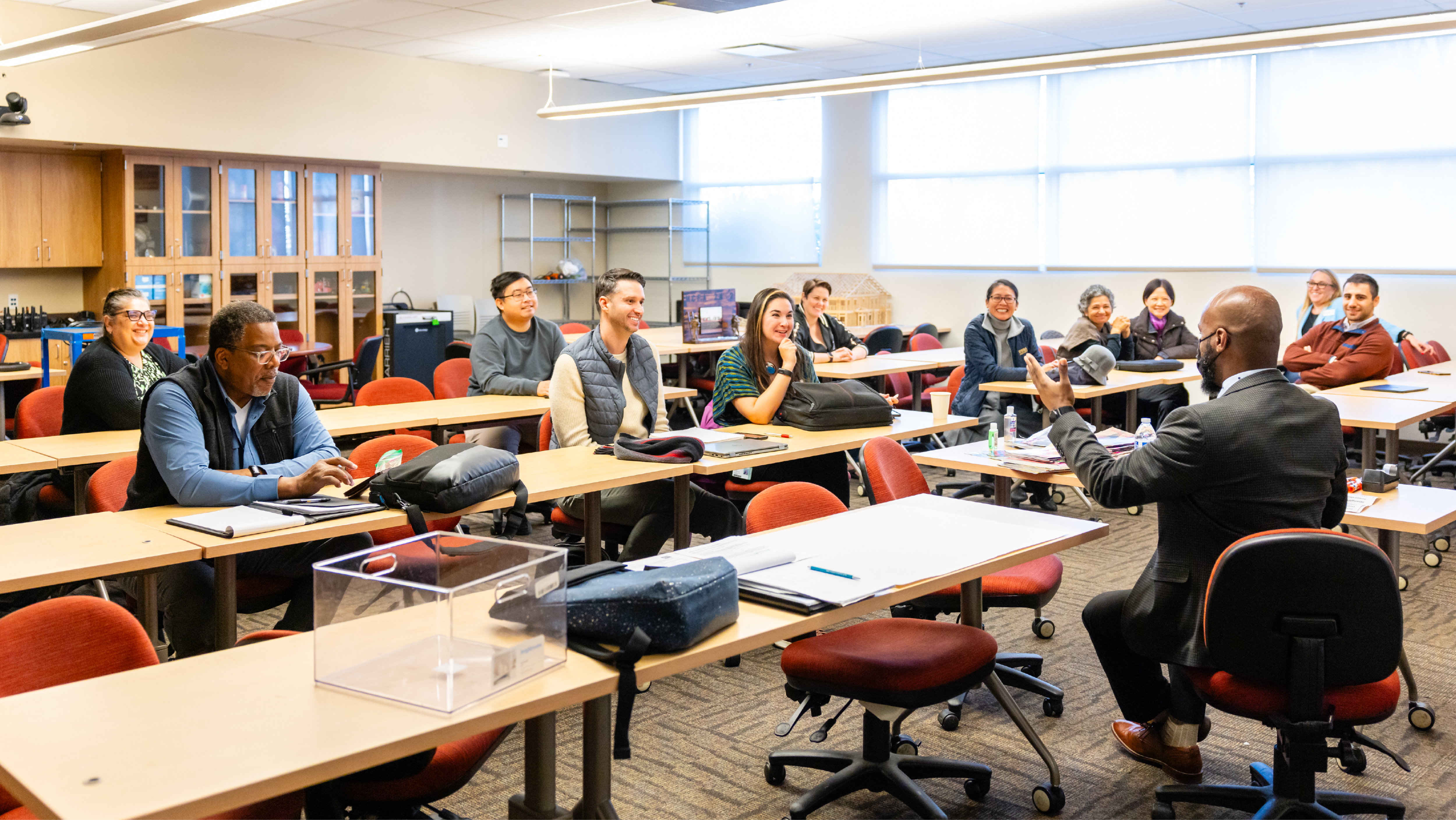 A instructor speaking to a diverse group of adult students seated at desks in a bright classroom.