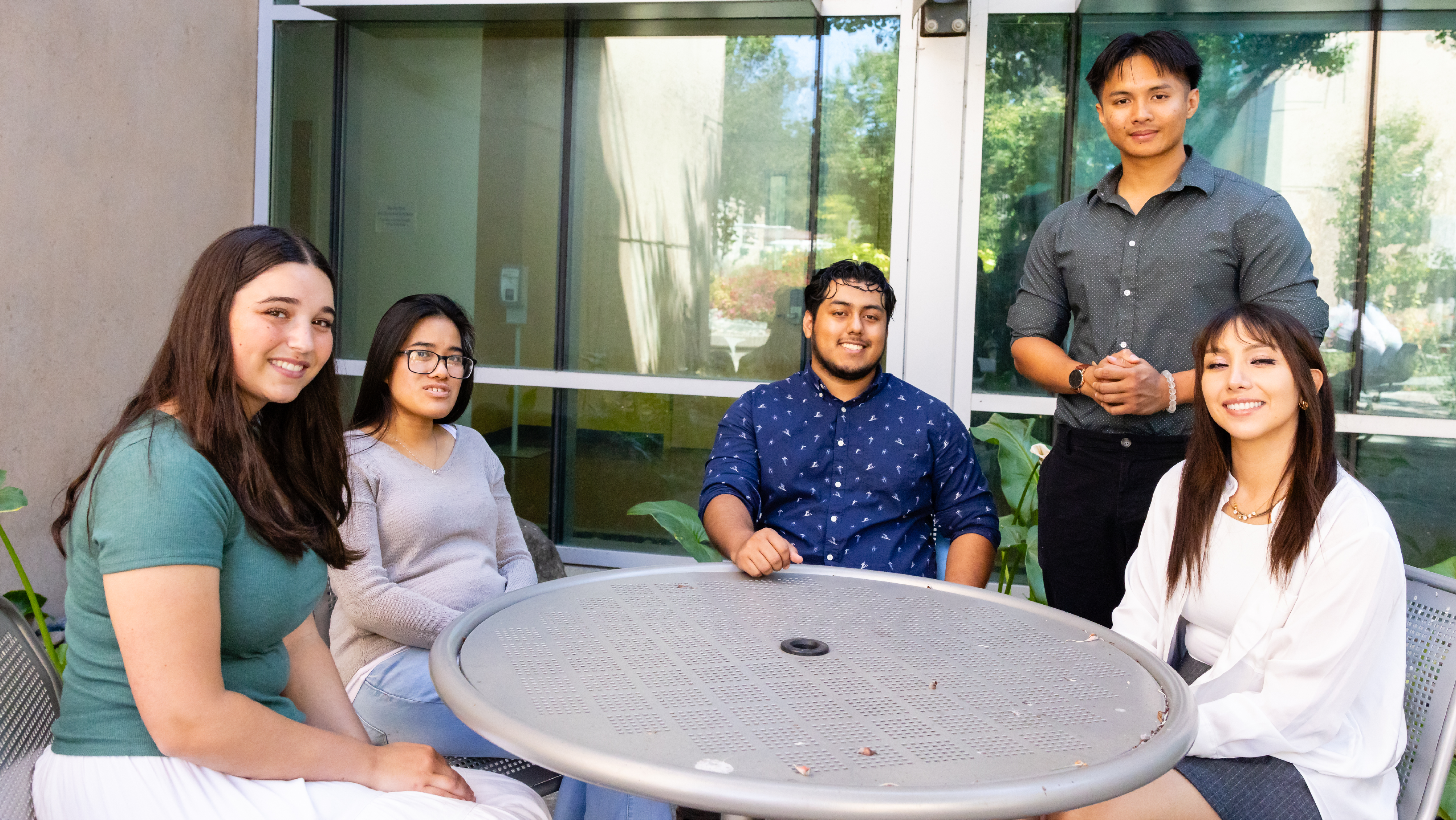 Five smiling students posing together around a circular outdoor table on a campus patio.