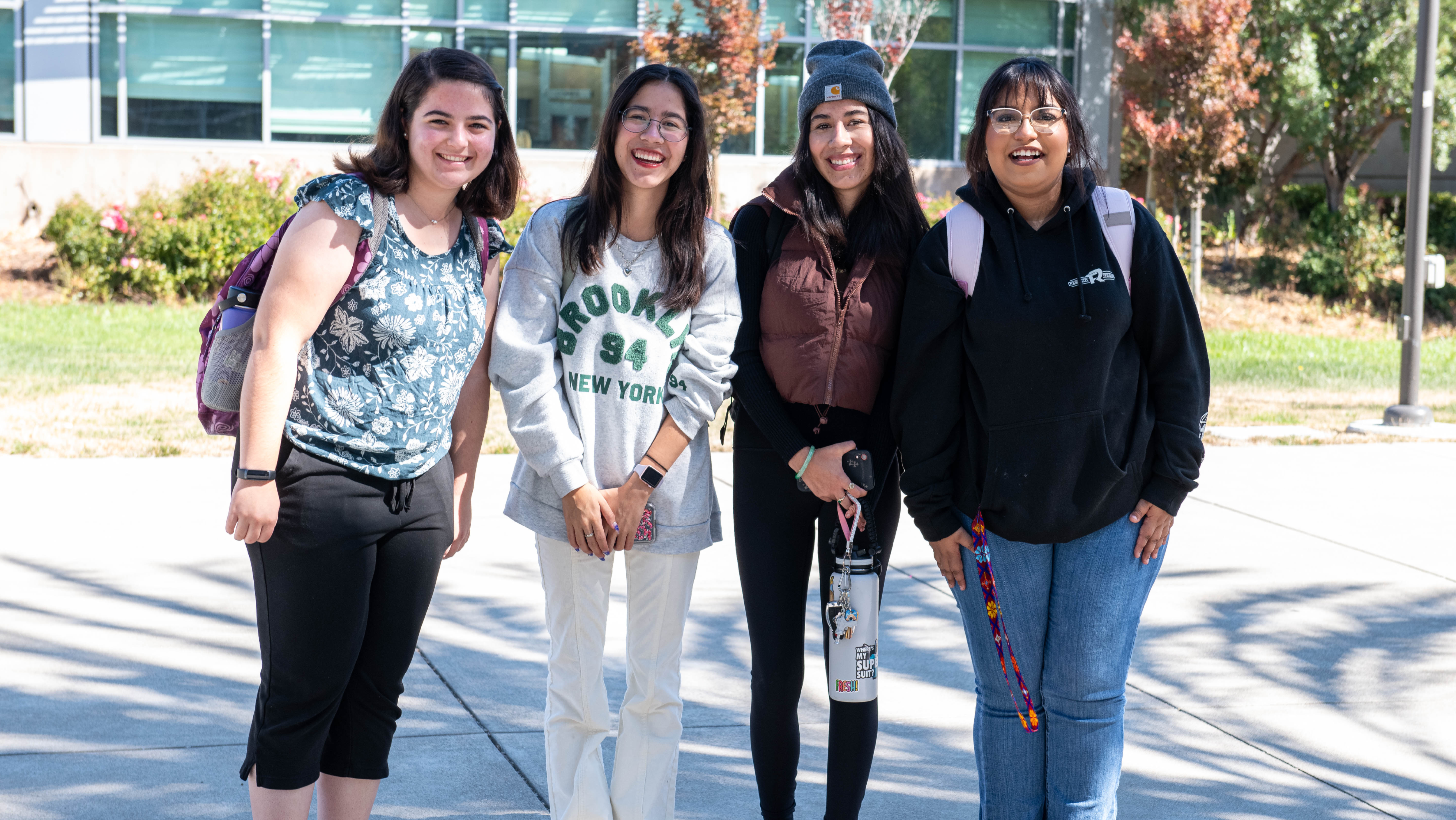 Four female students standing together outdoors on a sunny campus pathway, smiling at the camera.