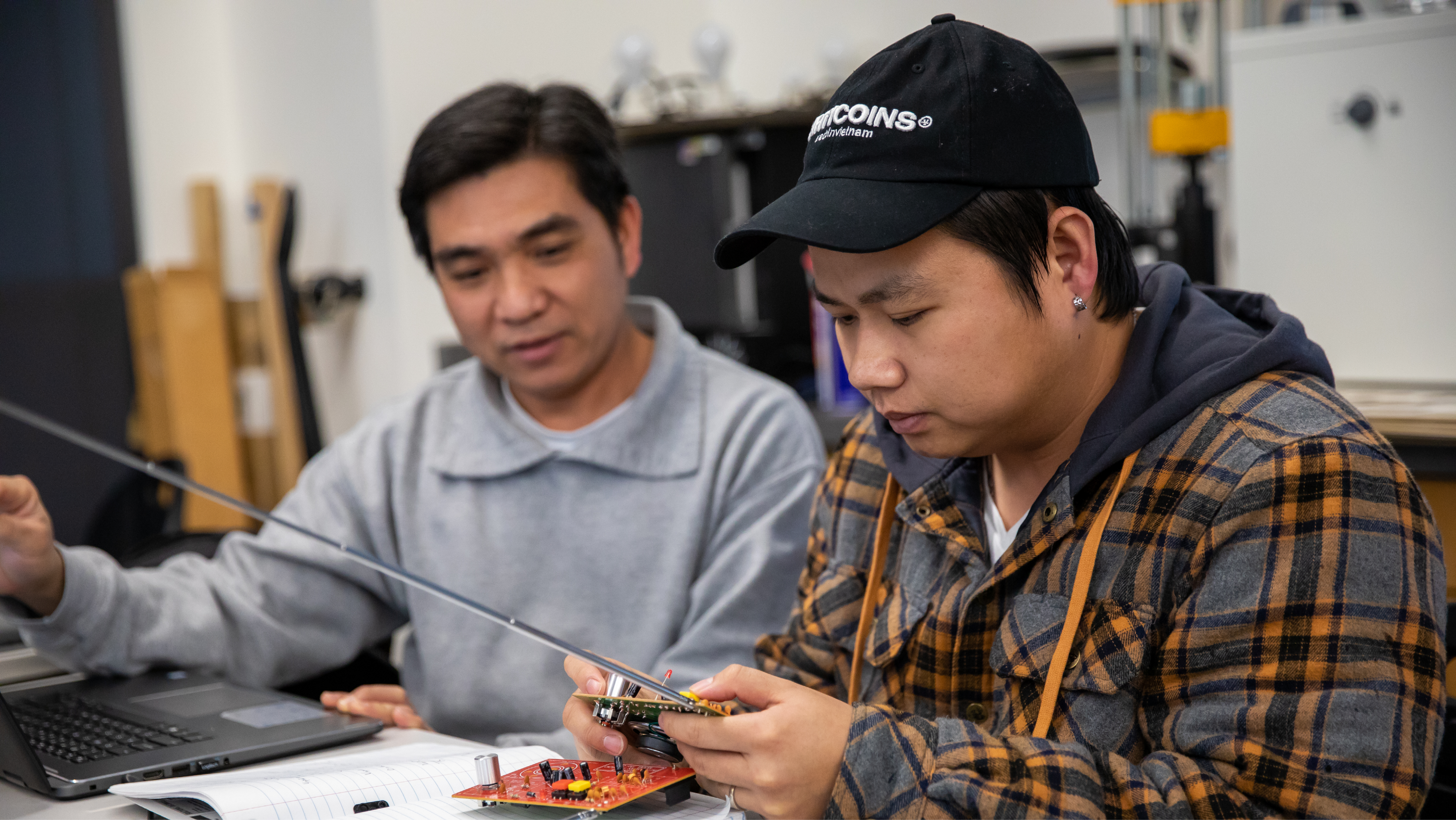Two men in a lab setting closely examining a circuit board and electronic component.