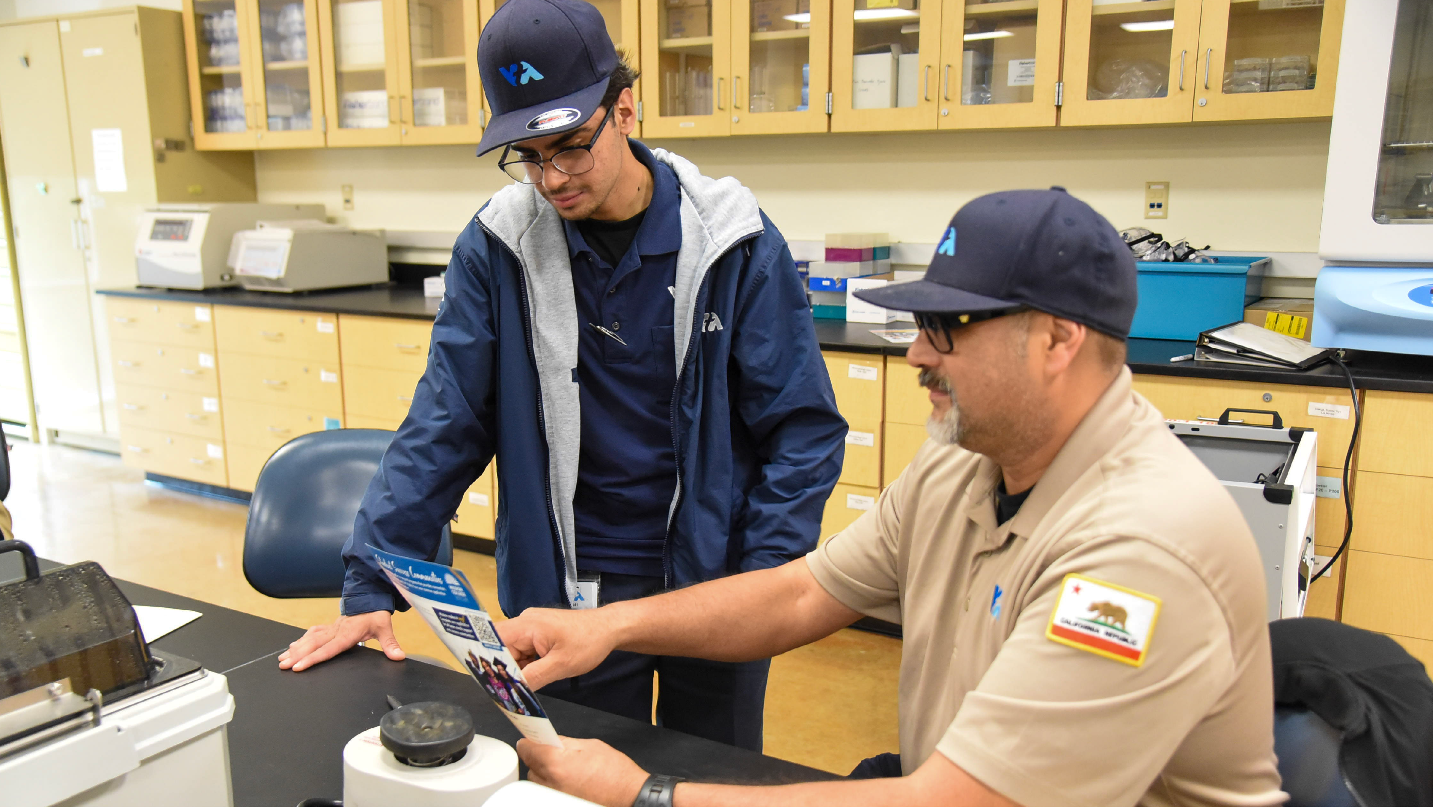 Two men wearing VTA caps and branded apparel reviewing printed program materials together at a lab table.