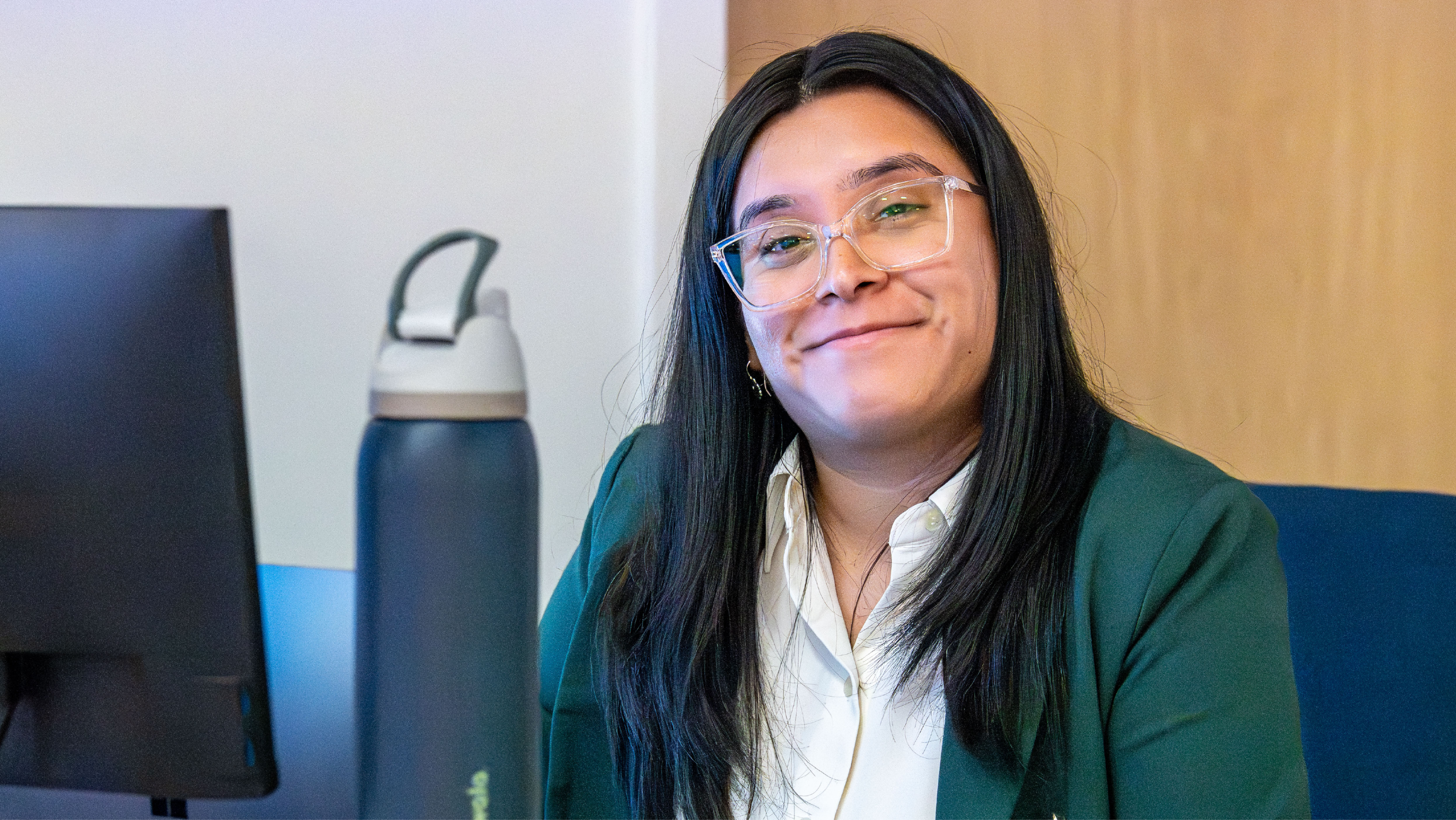 A smiling female professional with glasses seated at a office desk in front of a computer monitor.
