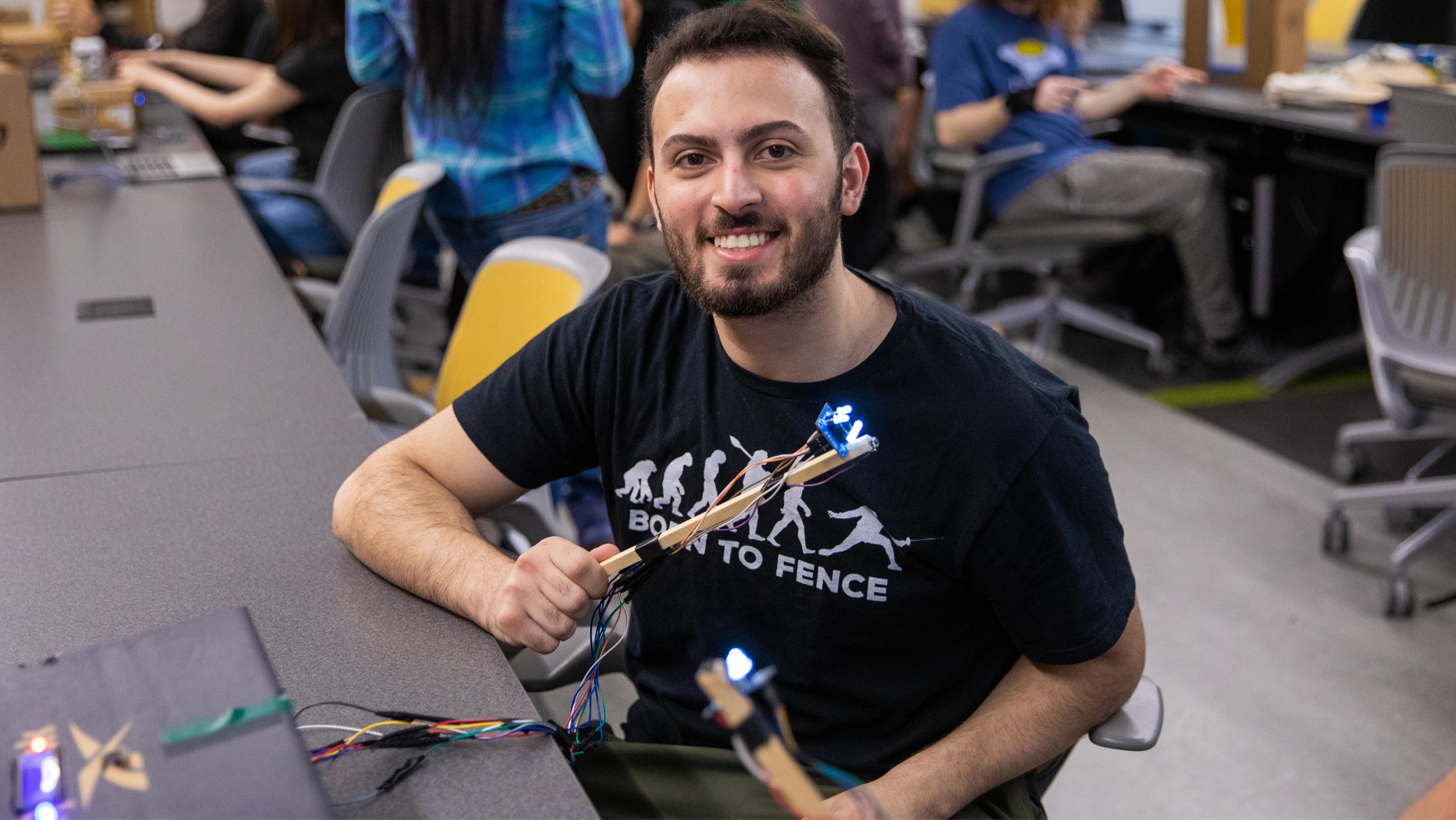 A male student sitting at a lab bench smiling while demonstrating an electronic device with small lit LED lights.