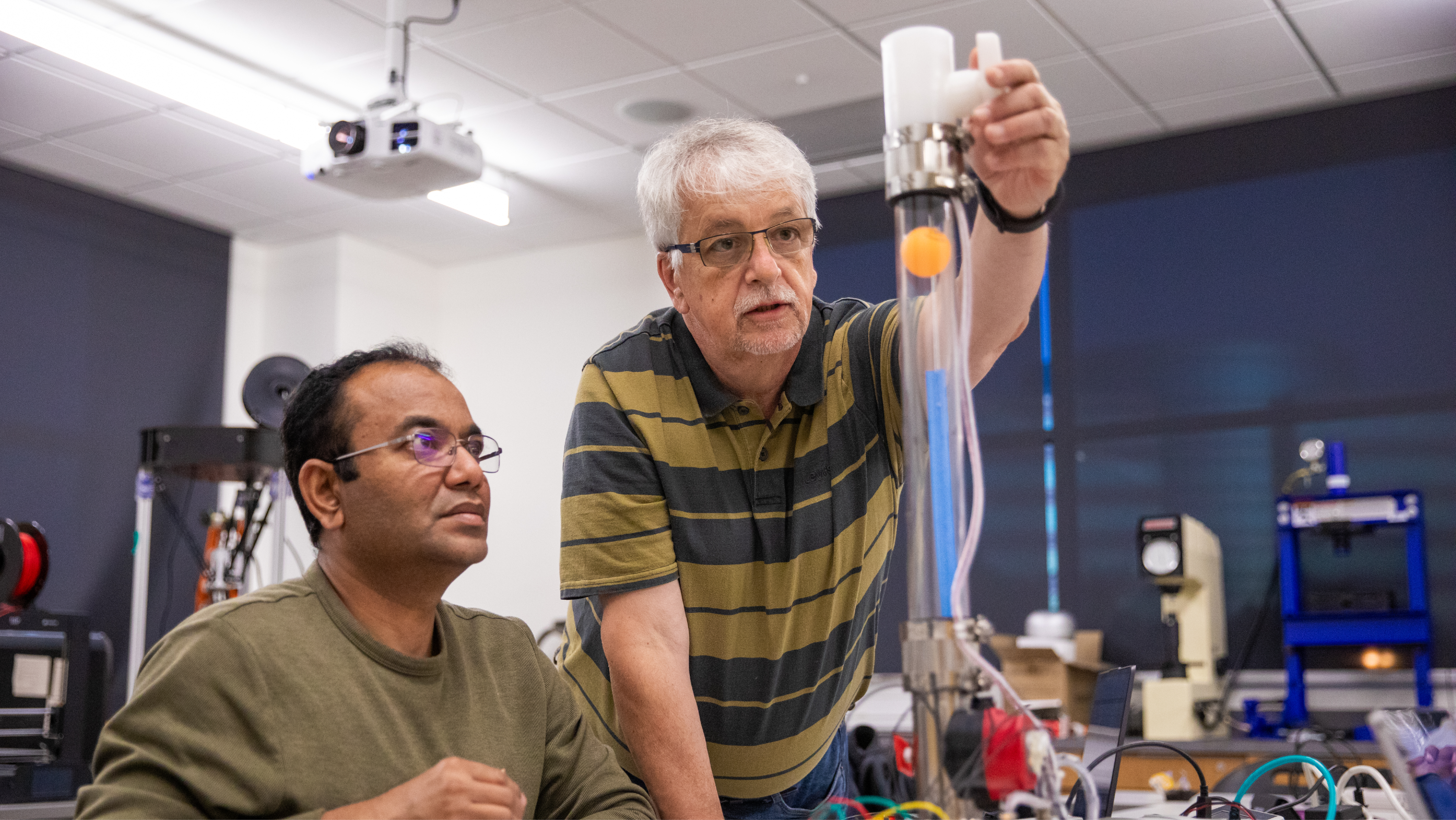 An instructor and a student in a lab setting observing a clear vertical tube mechanism.