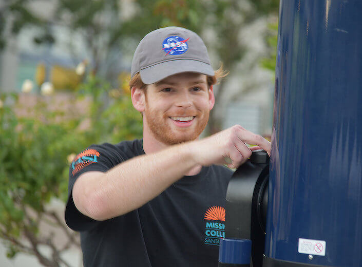 Redhaired male college student wears a blue NASA hat as he works with a telescope outside on a college campus.