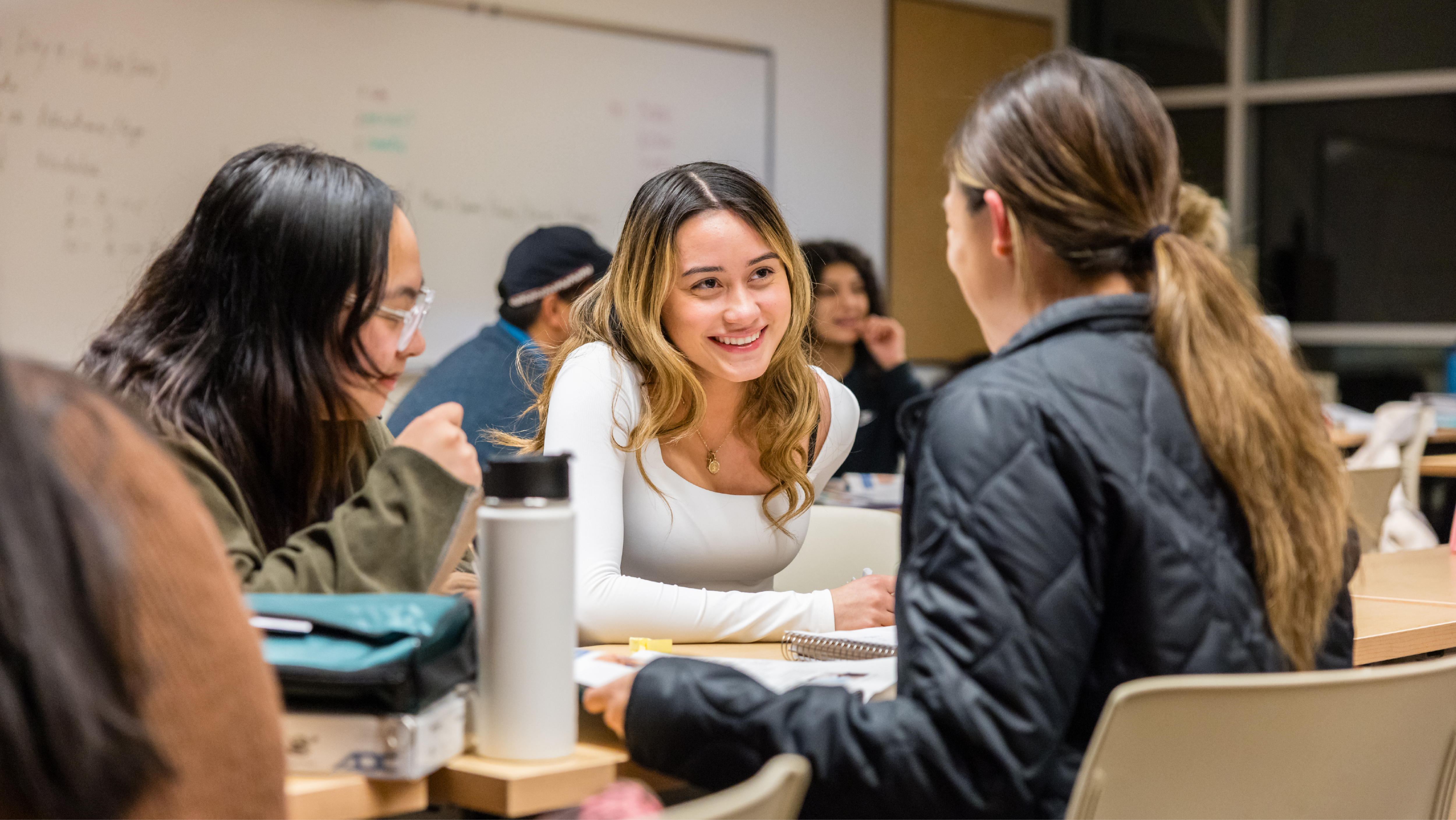 Three female students smiling and conversing while sitting around a classroom desk.