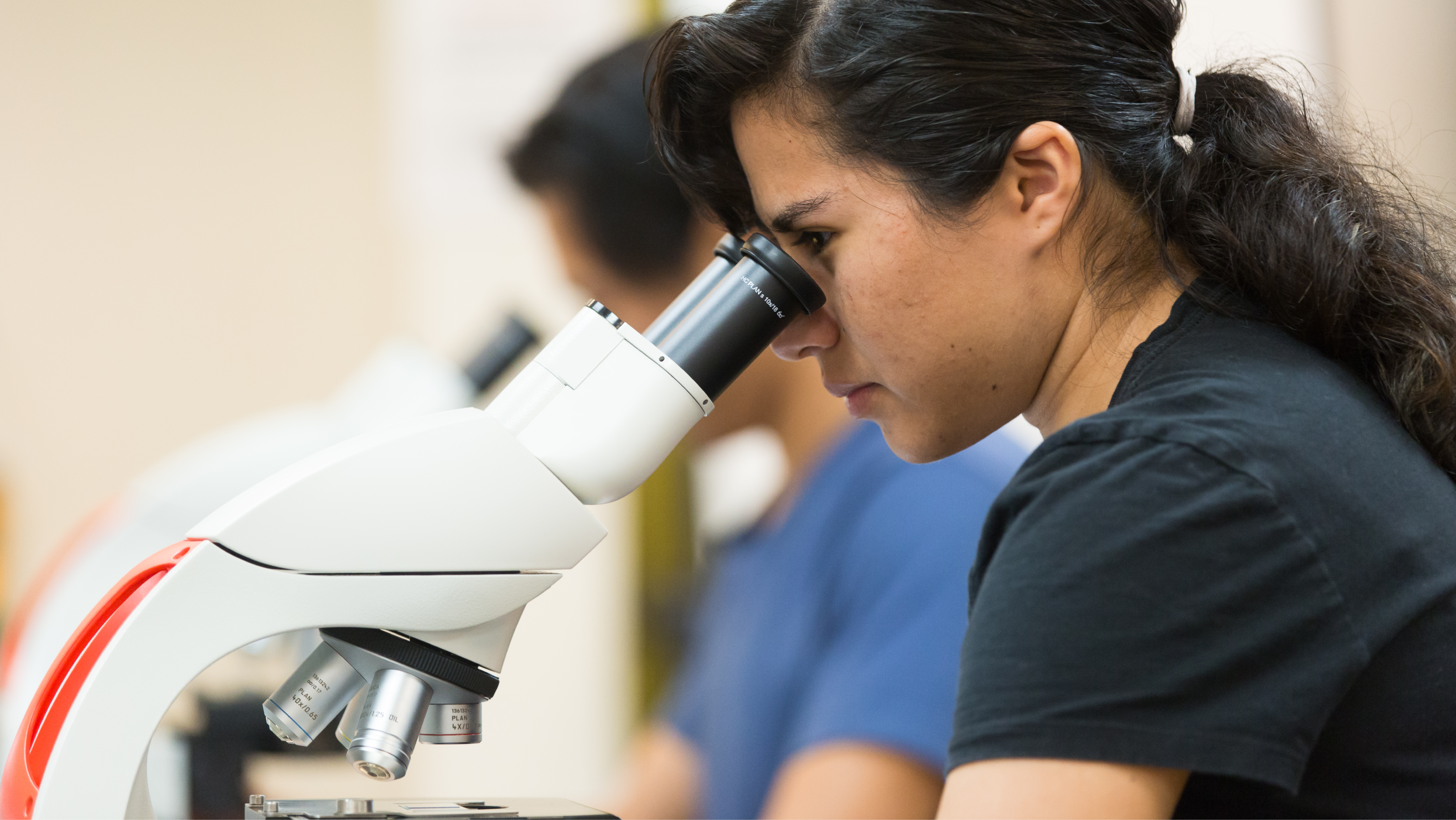 A female student looking closely through a binocular lab microscope during a class session.