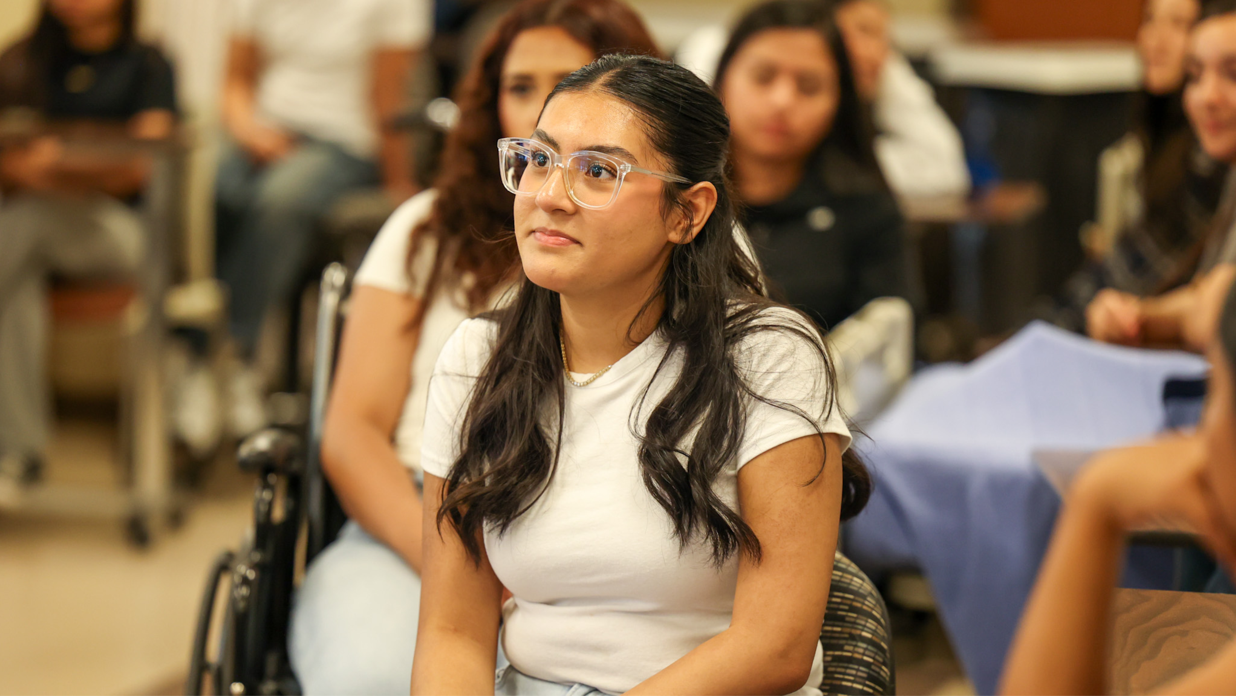 A female student wearing clear-framed glasses listening attentively during a classroom lecture.