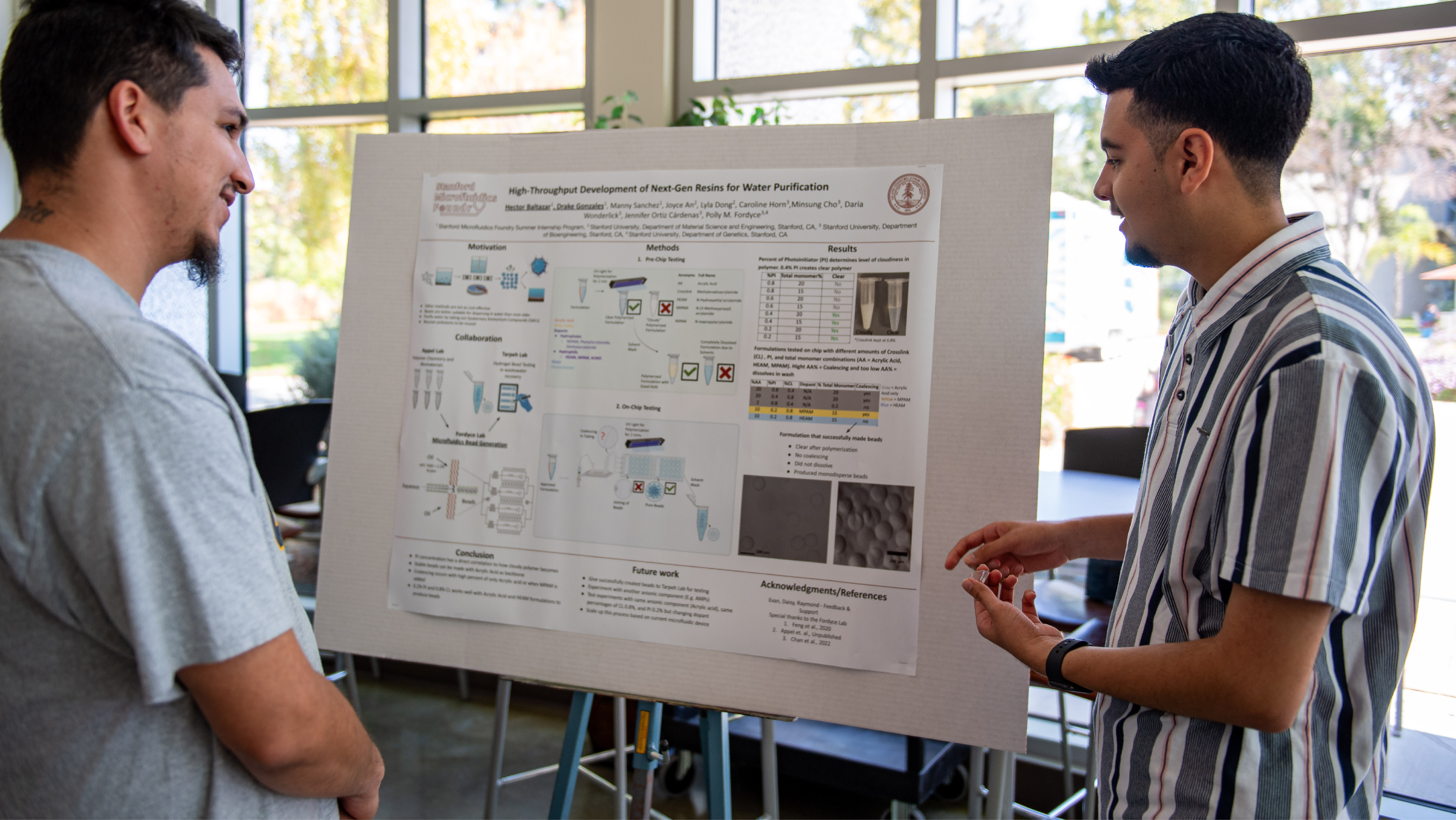 Two male students standing next to a poster presentation board and discussing water purification research.