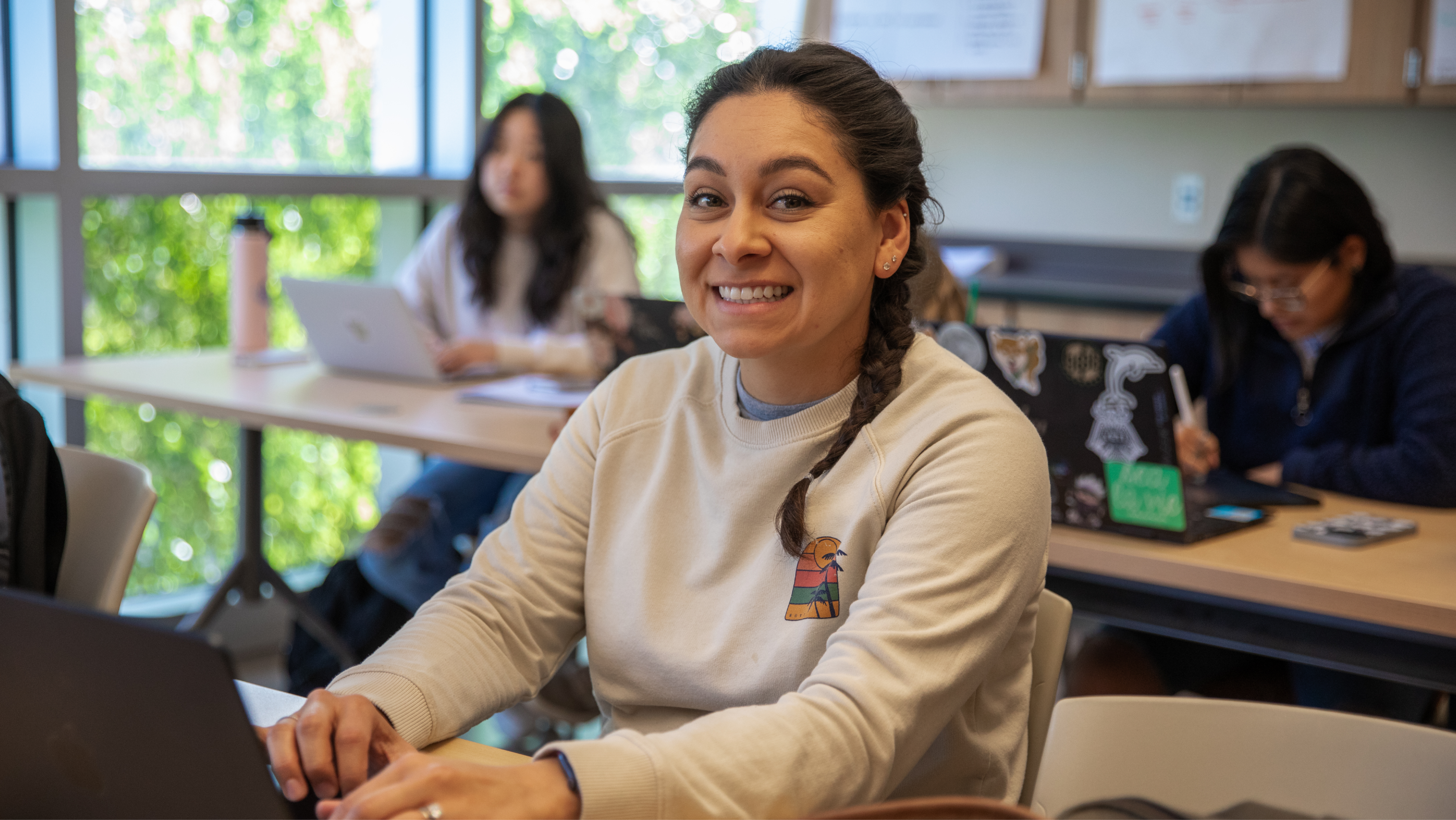 A smiling female student seated at a desk typing on a laptop computer in a bright classroom.