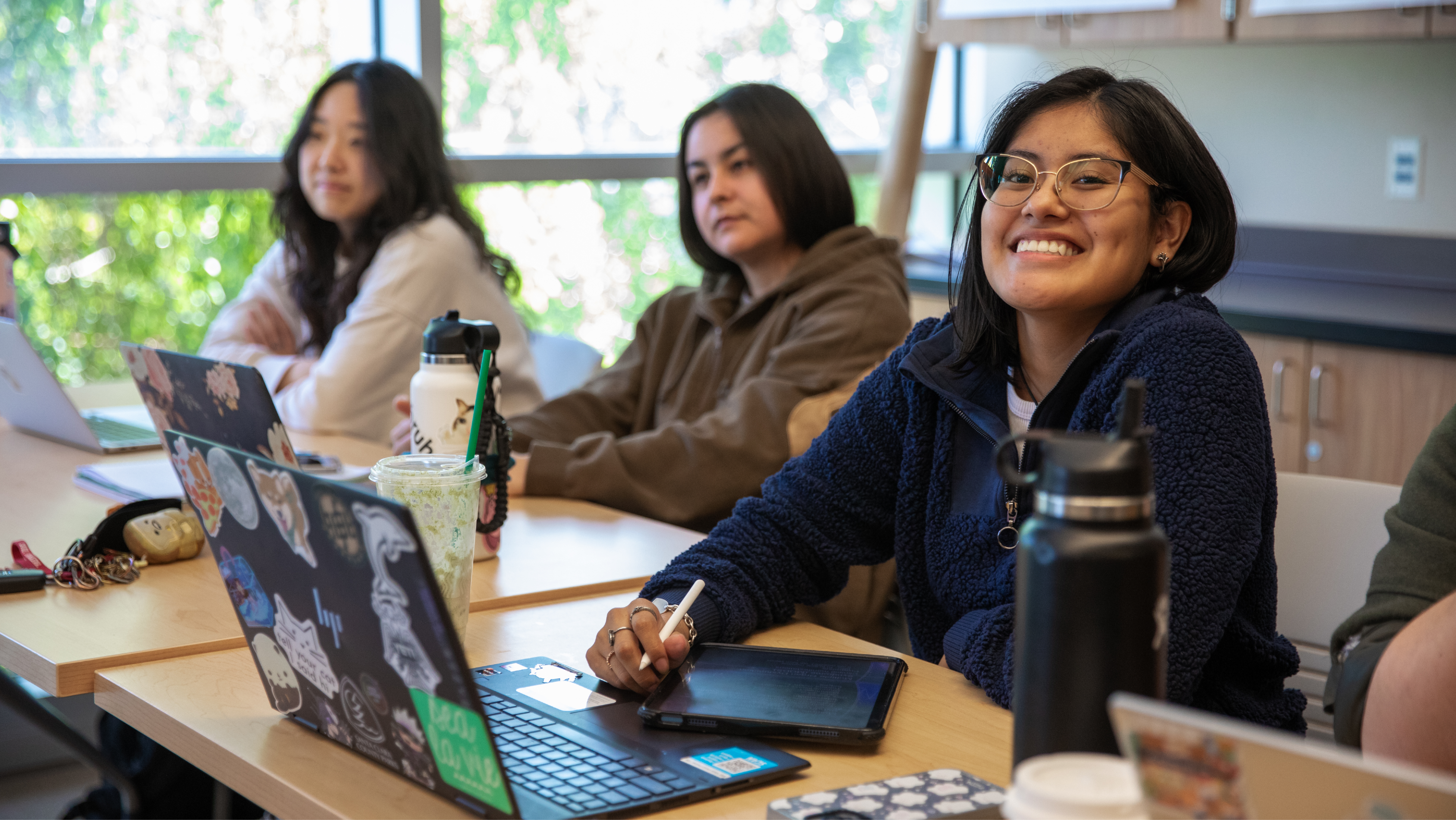 A female student in glasses smiling while taking notes on a tablet computer at a study table.