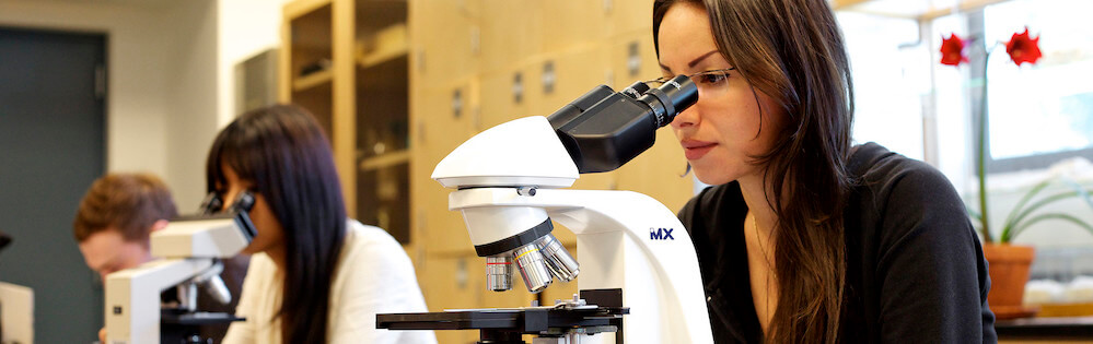 A young woman with straight brown hair past her shoulders looks into a microscope to examine a sample in Biology class.
