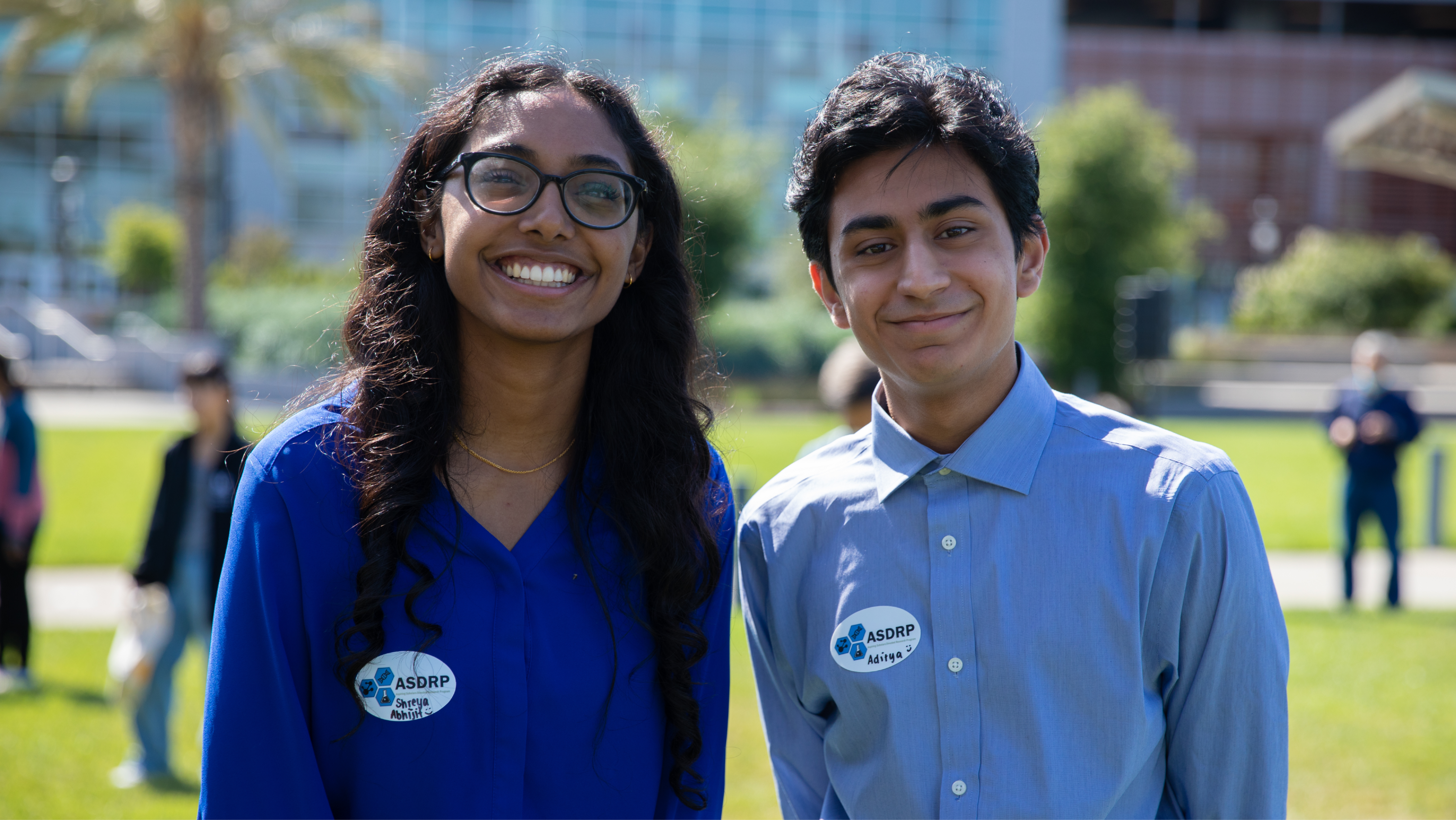 Two smiling students wearing name badges and professional attire standing outdoors on campus.