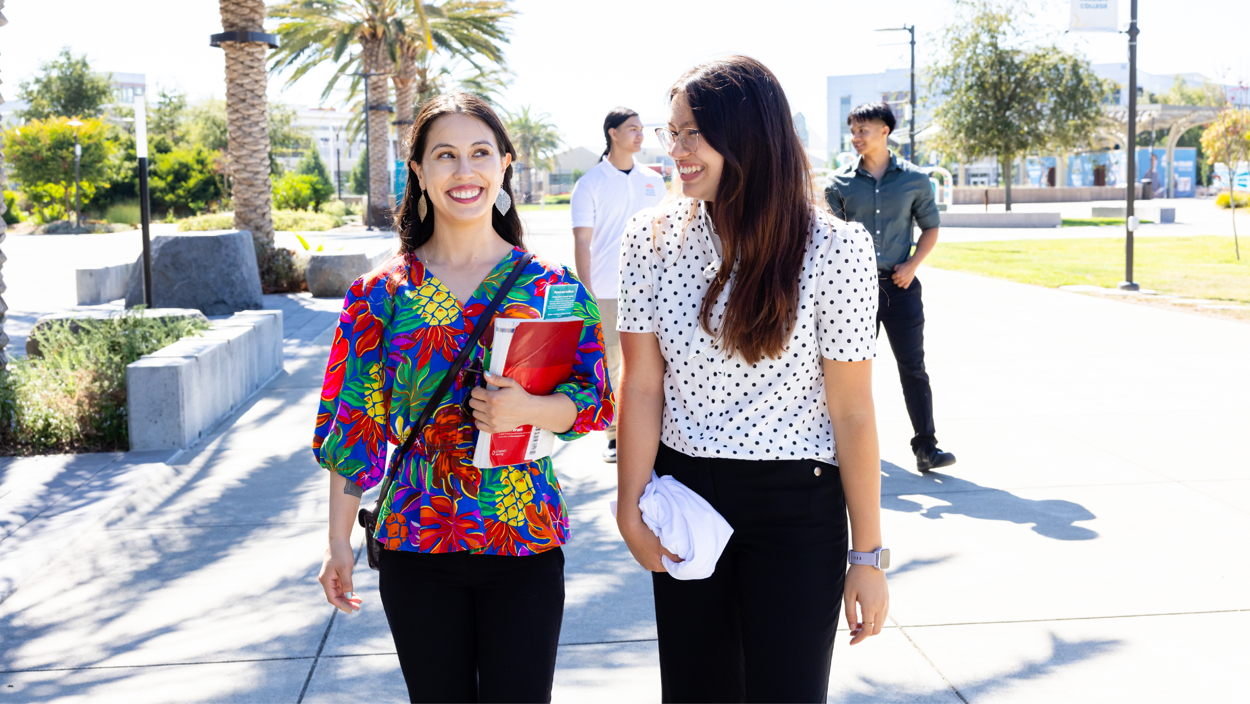 Two smiling female students carrying folders and notebooks while walking along a sunny campus walkway.