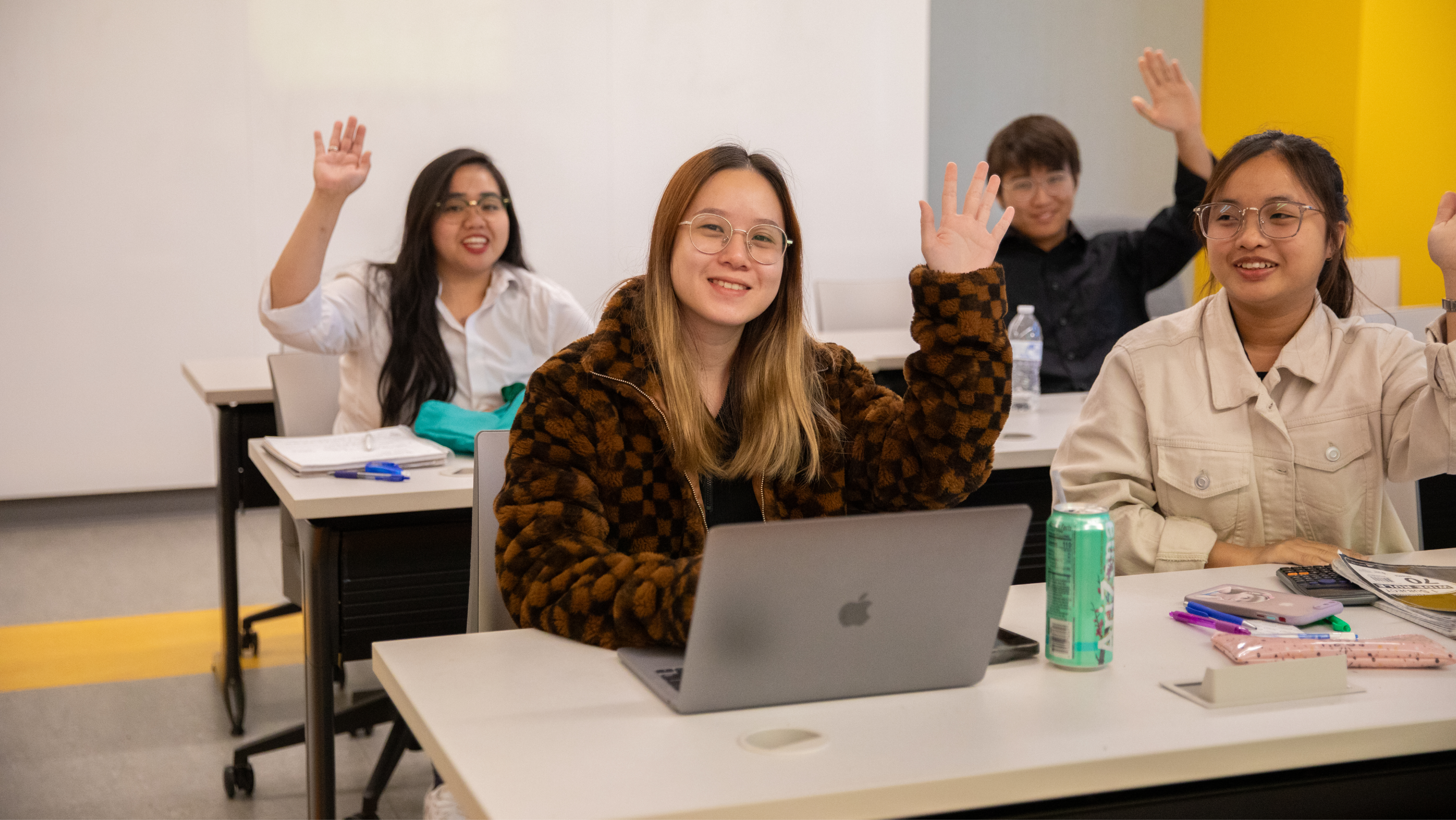 Four students sitting at desks in a classroom with their hands raised to participate.