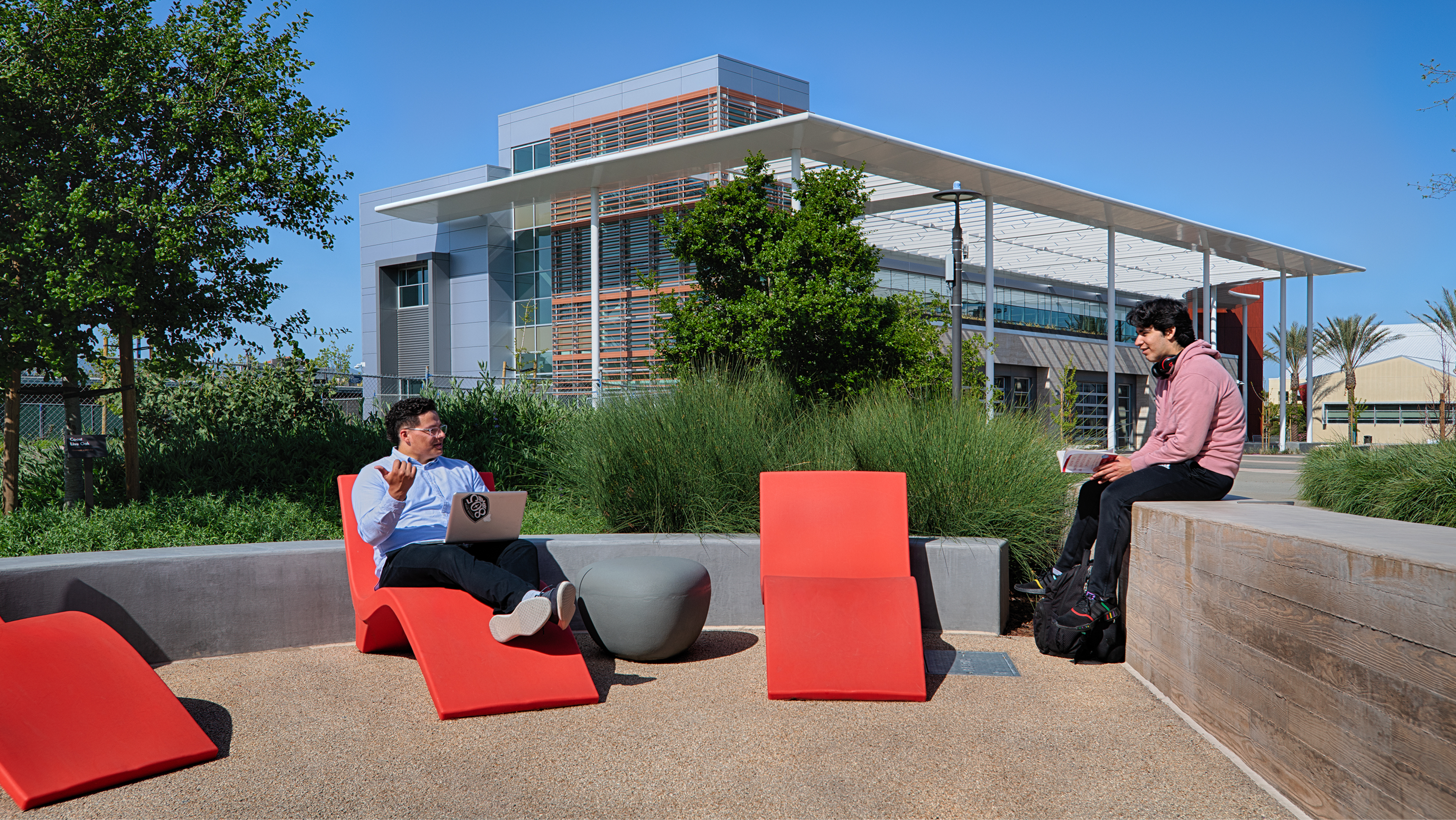 Two students studying outdoors on red modern lounge chairs in front of campus buildings.