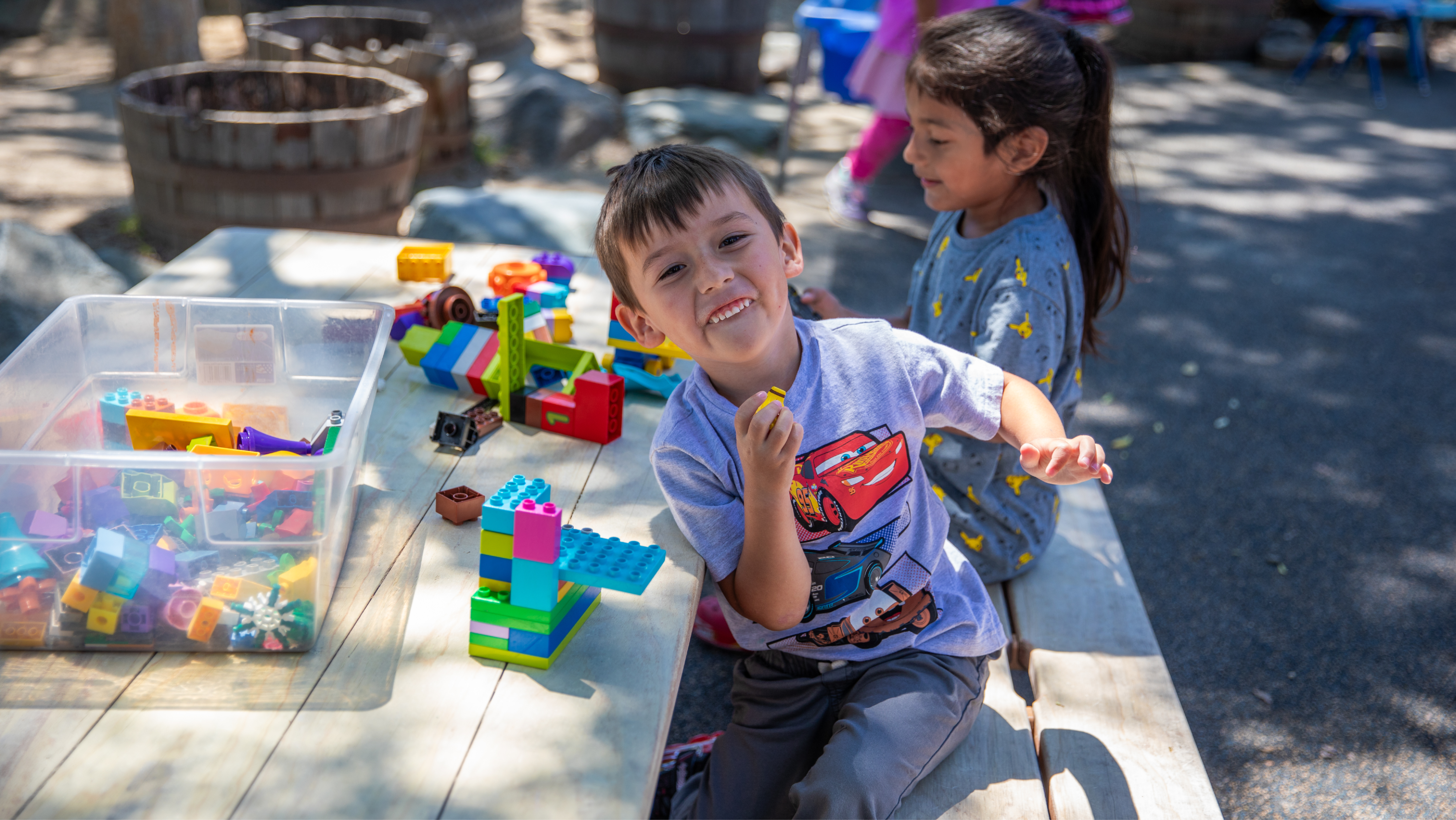 A young boy smiling at the camera while building a colorful tower out of plastic blocks at an outdoor wooden table.