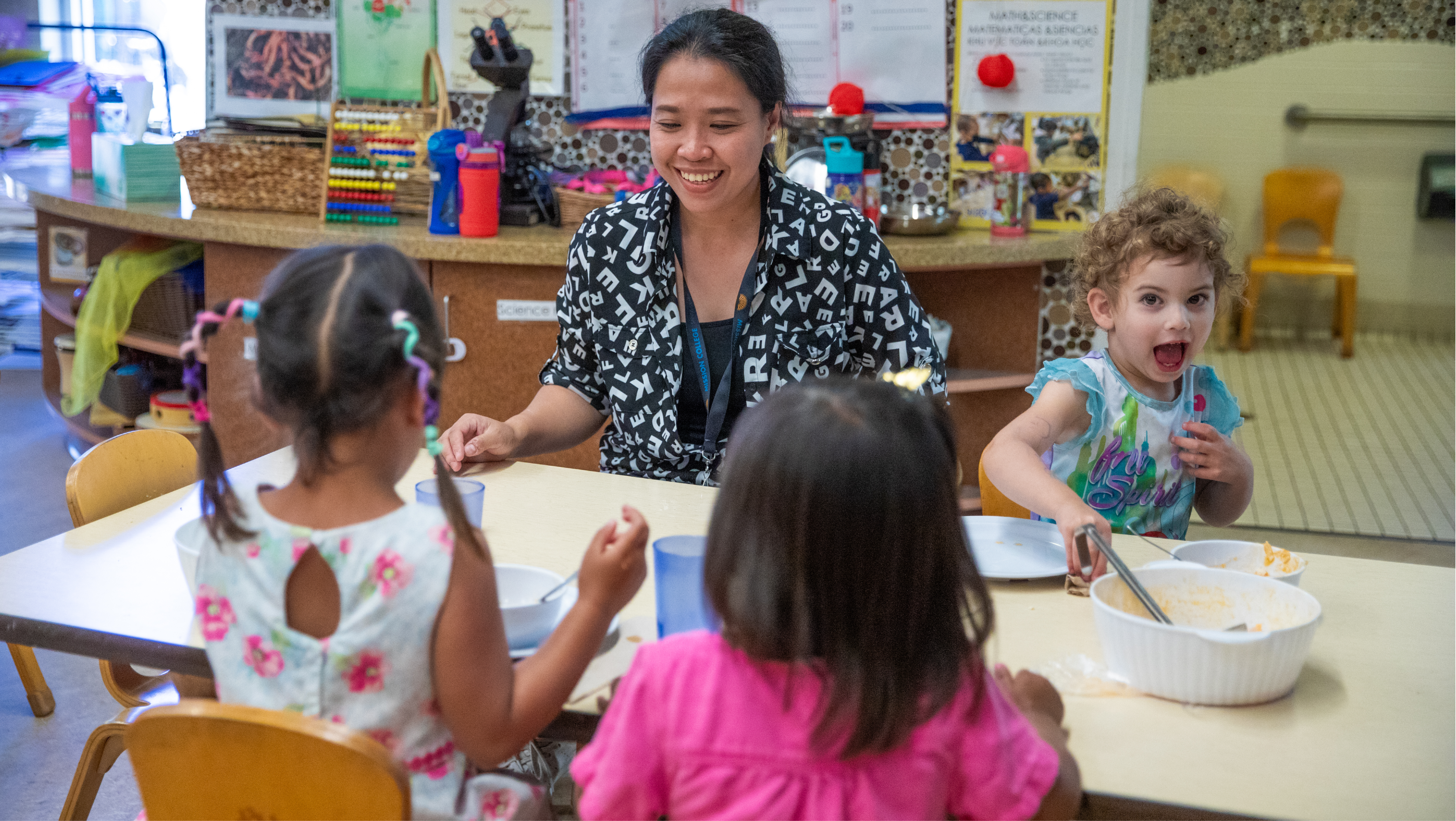 A female teacher smiling on a floor mat while holding a baby and supervising toddlers playing with colorful sensory balls.