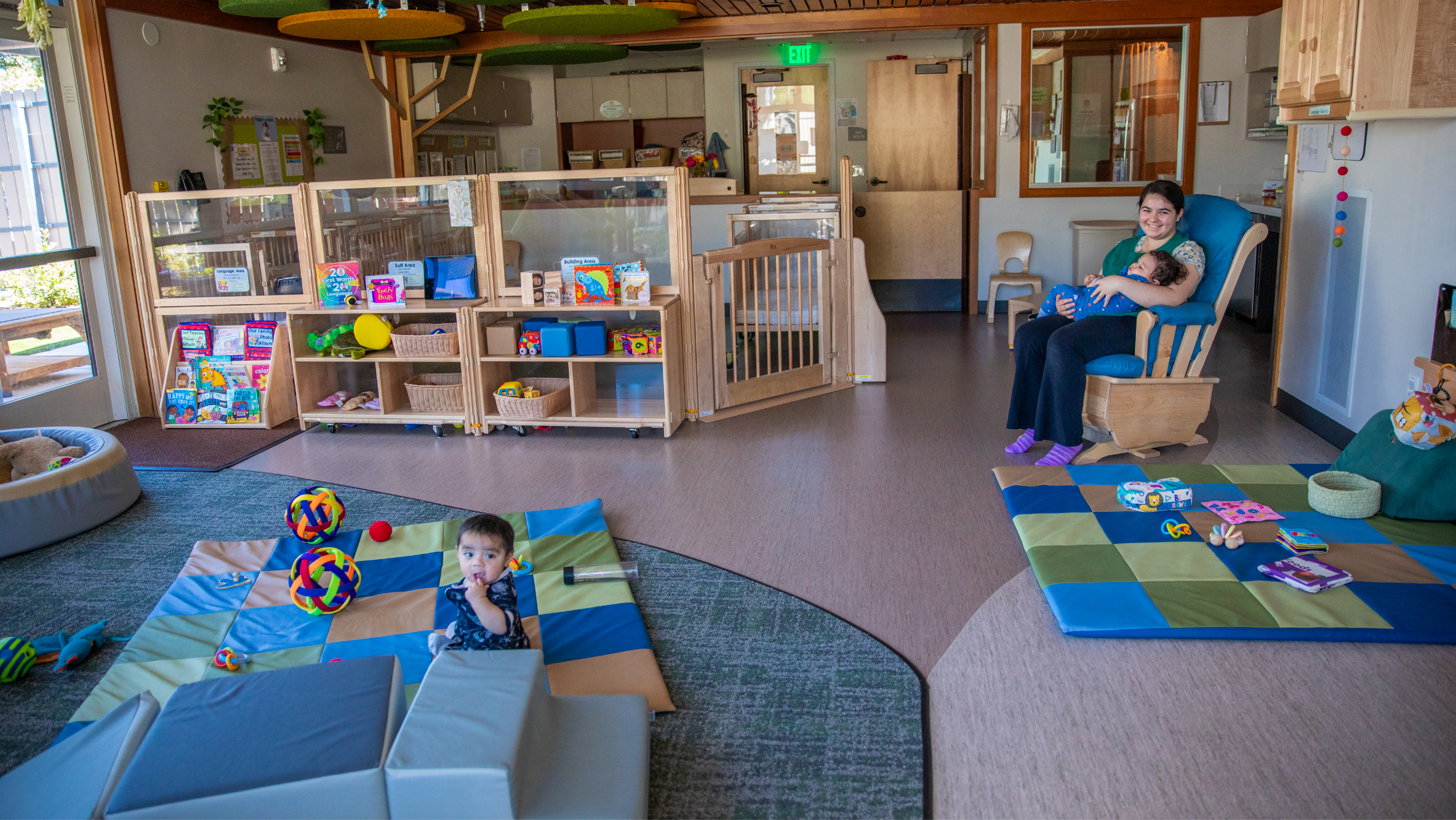 A wide view of a modern infant and toddler development center with soft floor mats, low bookshelves, and a teacher rocking a baby in a chair.