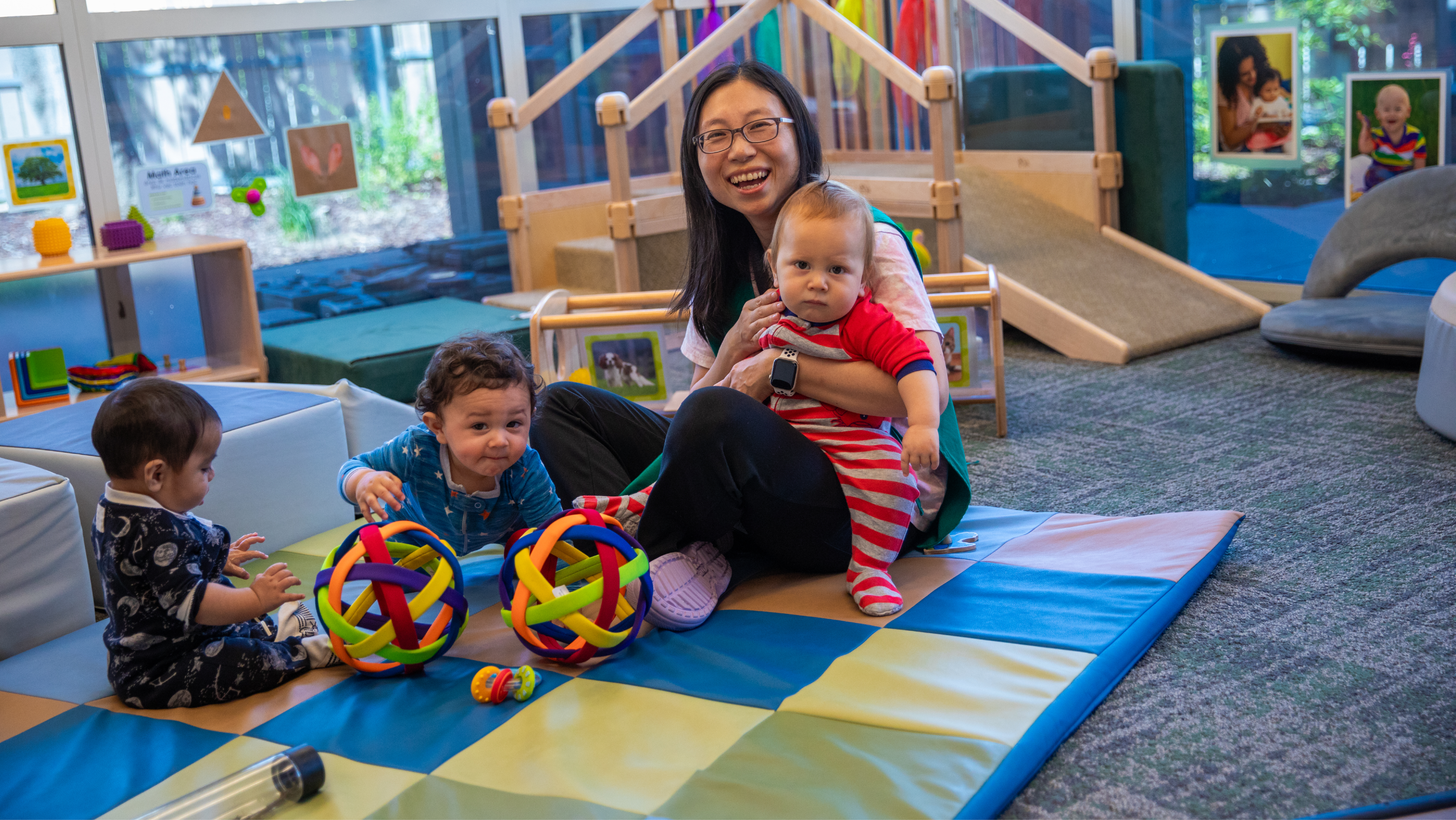 A female teacher smiling on a floor mat while holding a baby and supervising toddlers playing with colorful sensory balls.