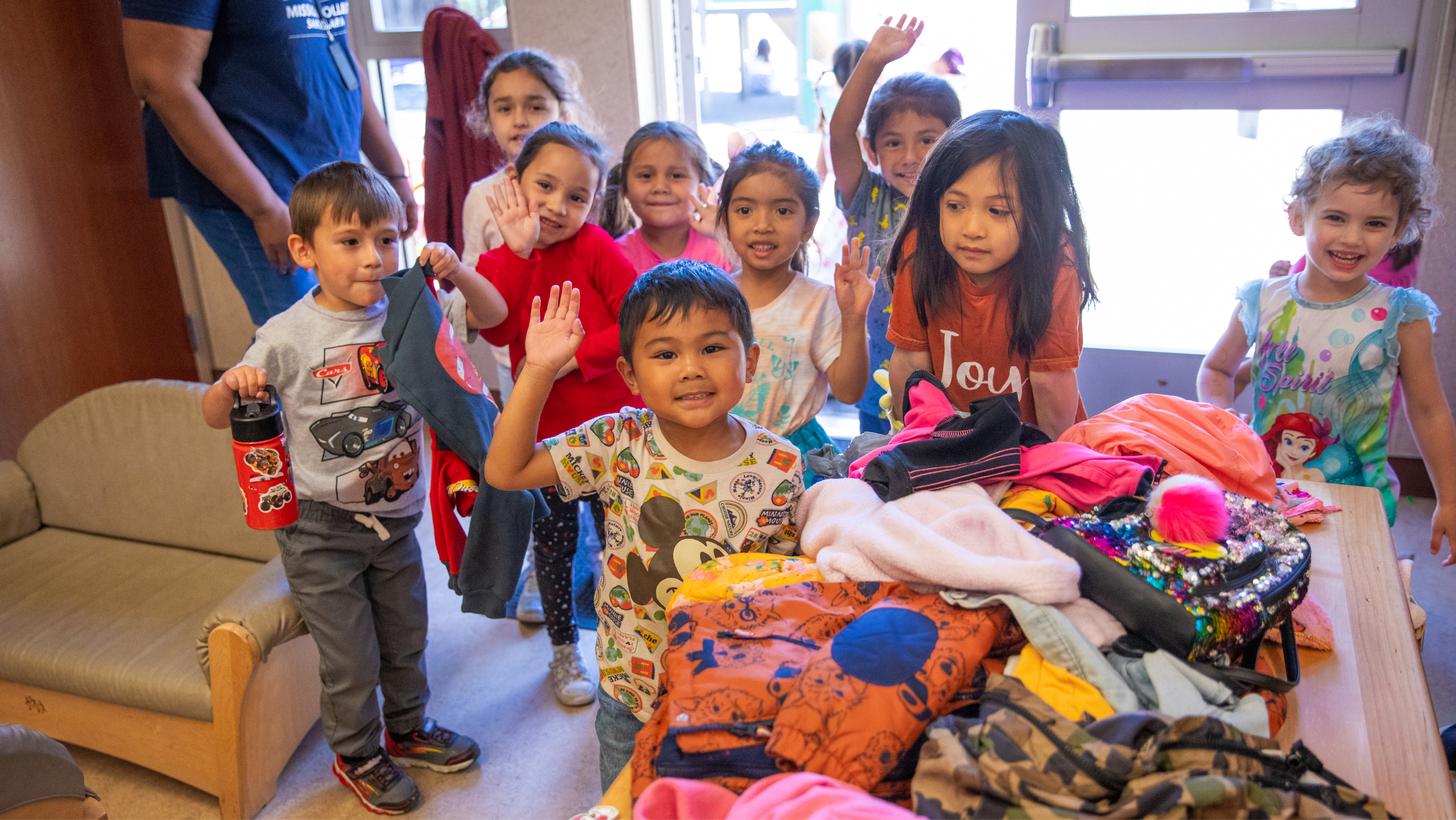 A group of young children waving happily at the camera while standing around a table piled with winter jackets and backpacks.