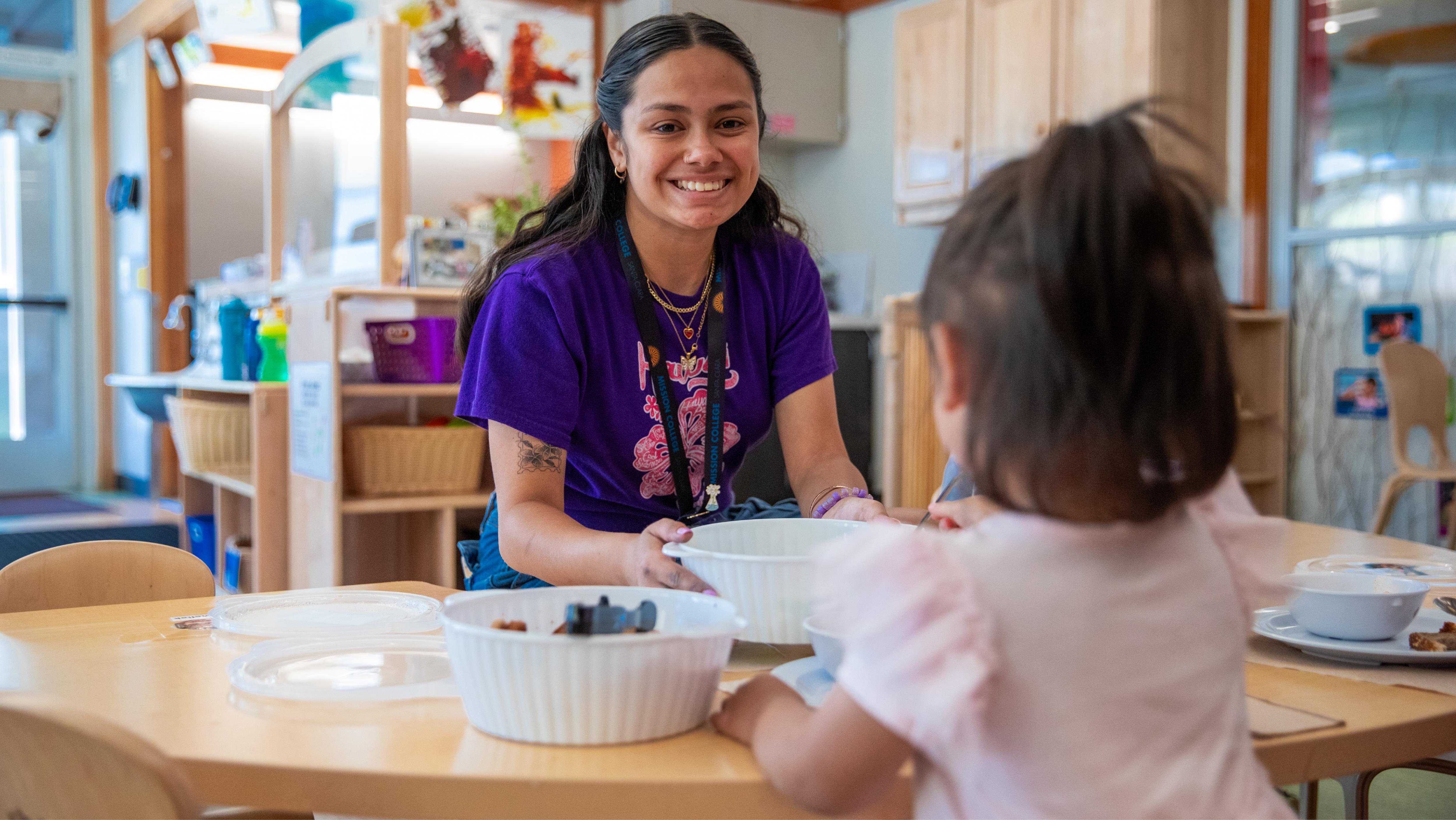 A teacher in a purple Mission College shirt smiling while assisting a toddler at a small dining table.