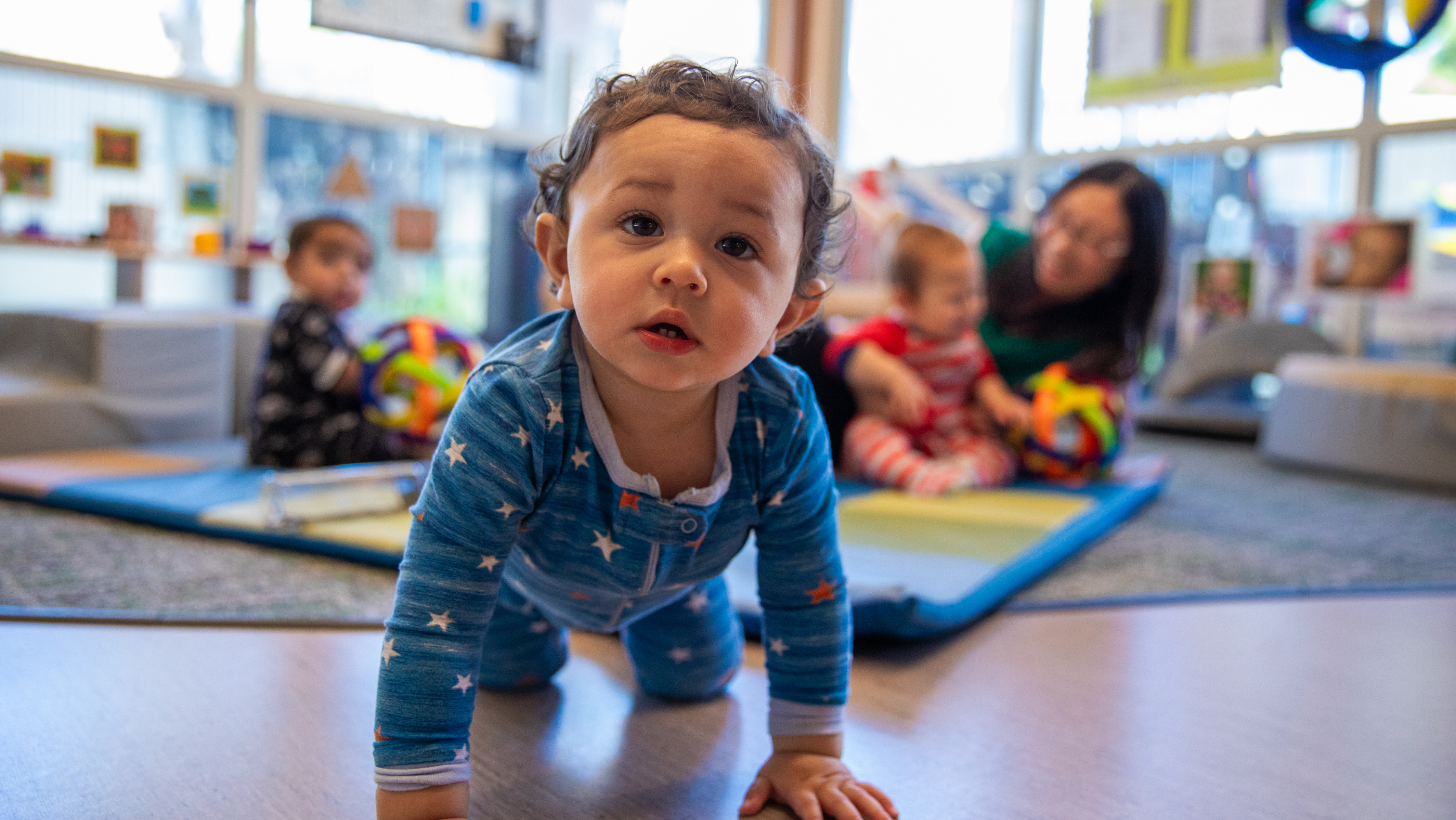 A toddler in blue star-patterned pajamas crawling forward on a play mat in a brightly lit infant care center.