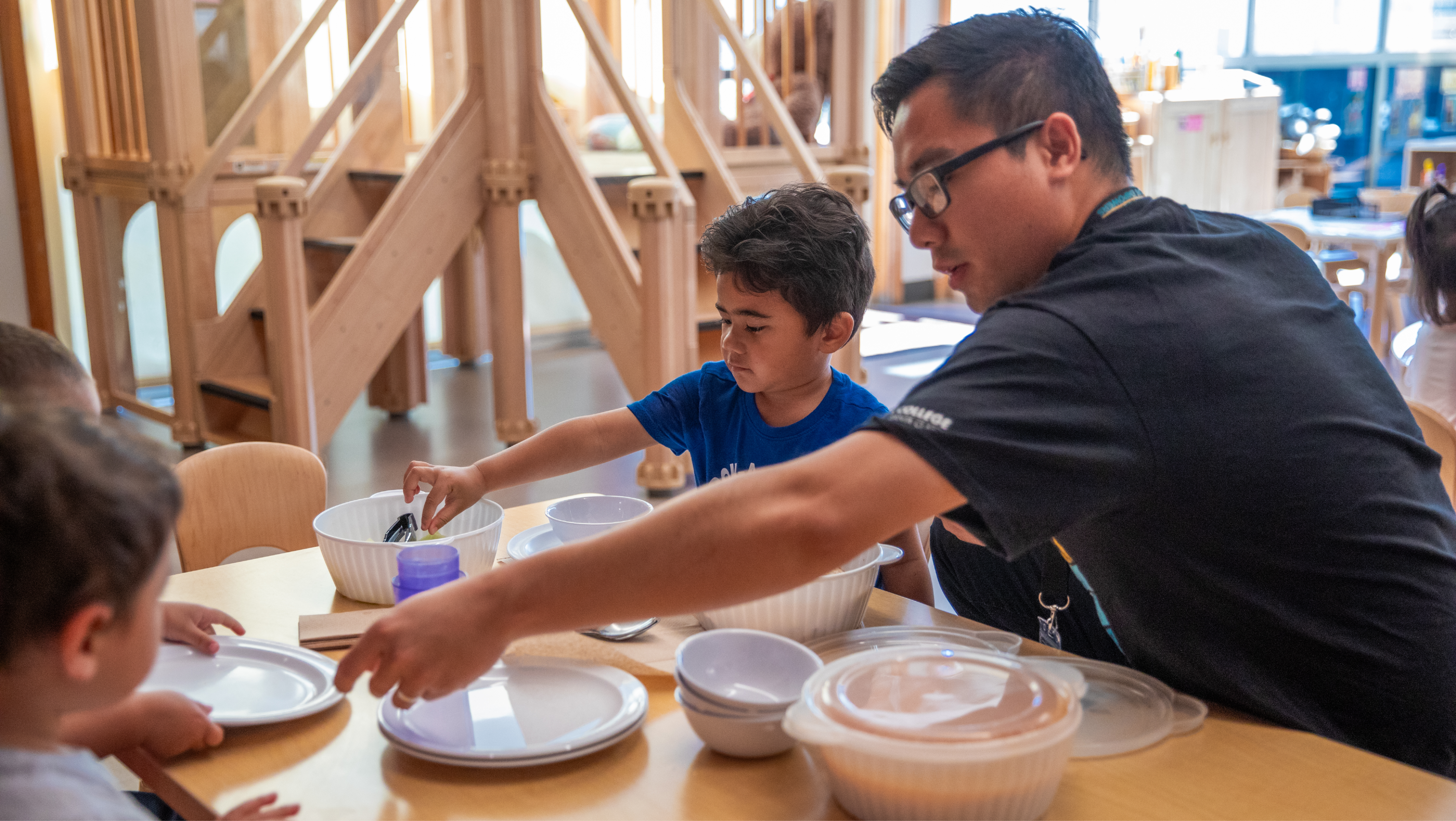 A male teacher helping a young child serve food at a wooden table in an early learning classroom.