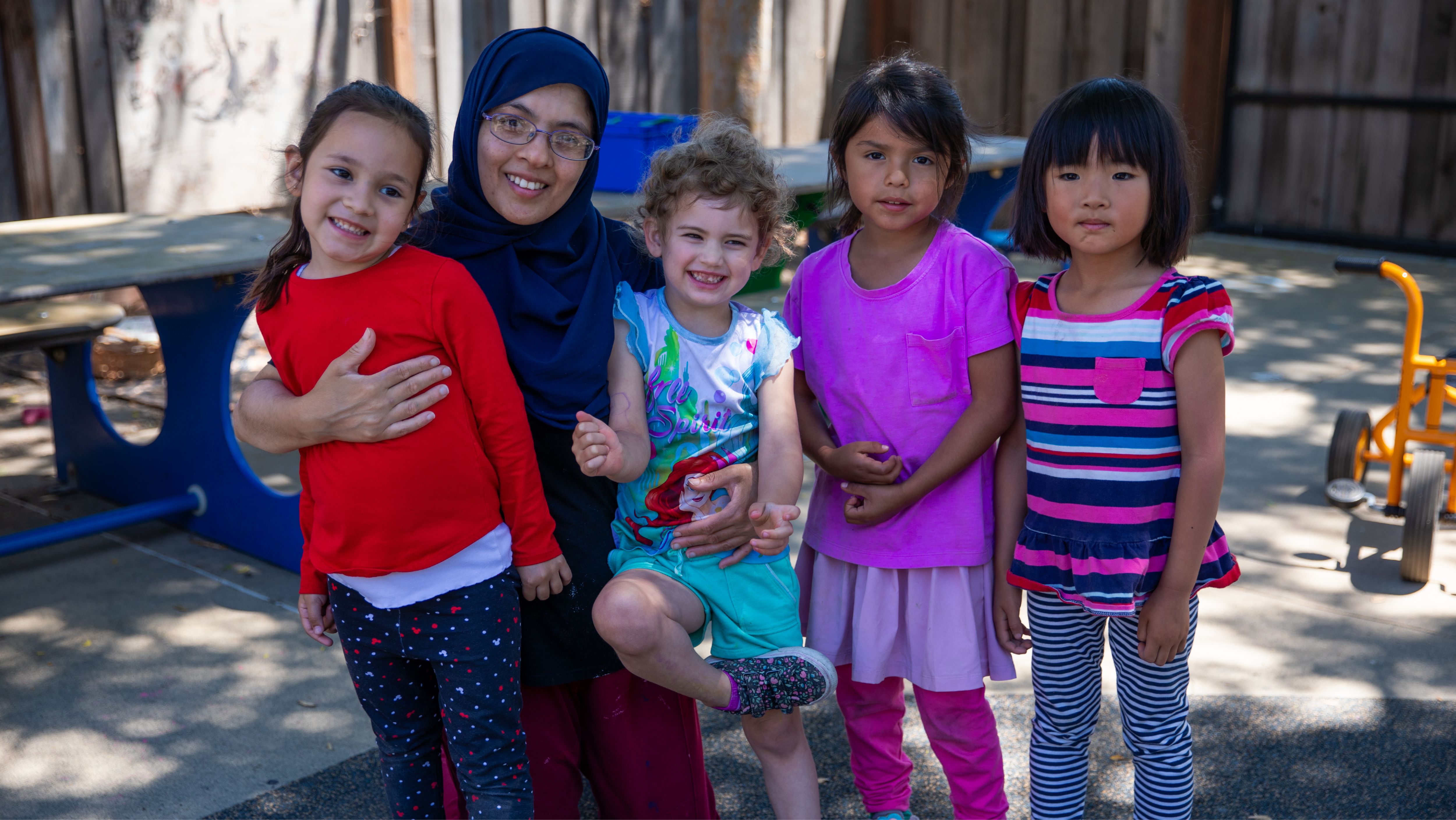 A female teacher wearing a navy hijab posing outdoors with four young children standing in a line beside her.