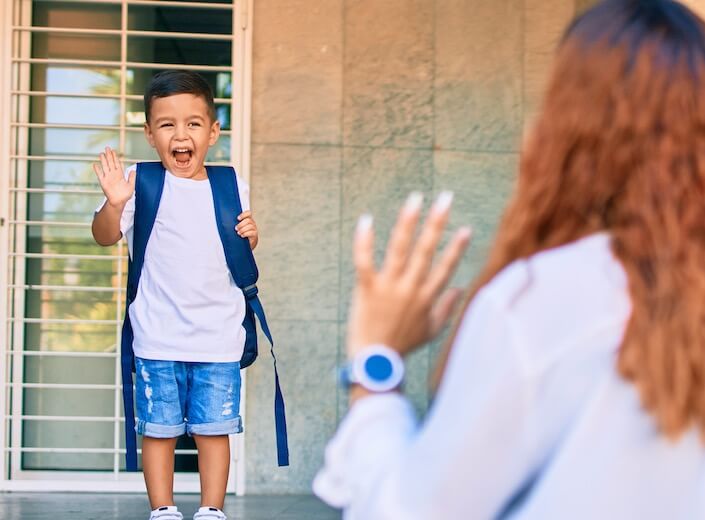 Preschool-aged boy of Latinx descent waves goodbye to his mom on the steps on a school.