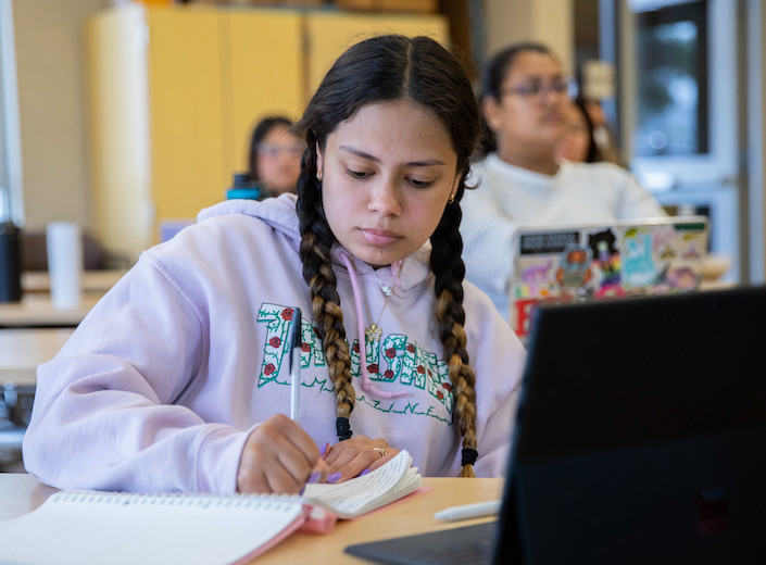 Early Childhood Development student writes in a notebook in her classroom. She is of Latinx descent and has thick dark braids and a nose ring.