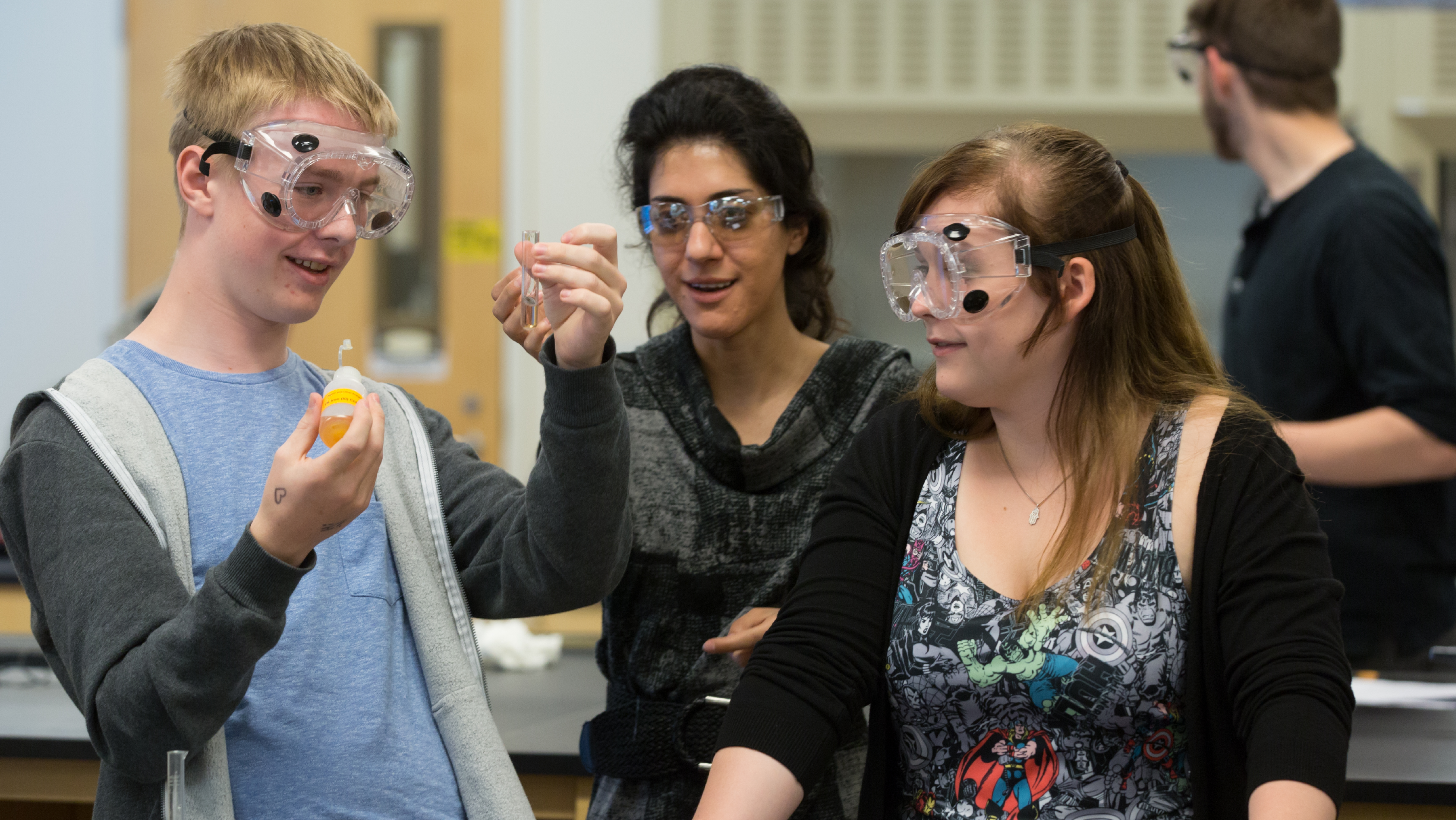 Four male students wearing safety goggles working in front of fume hoods and a periodic table in a lab.