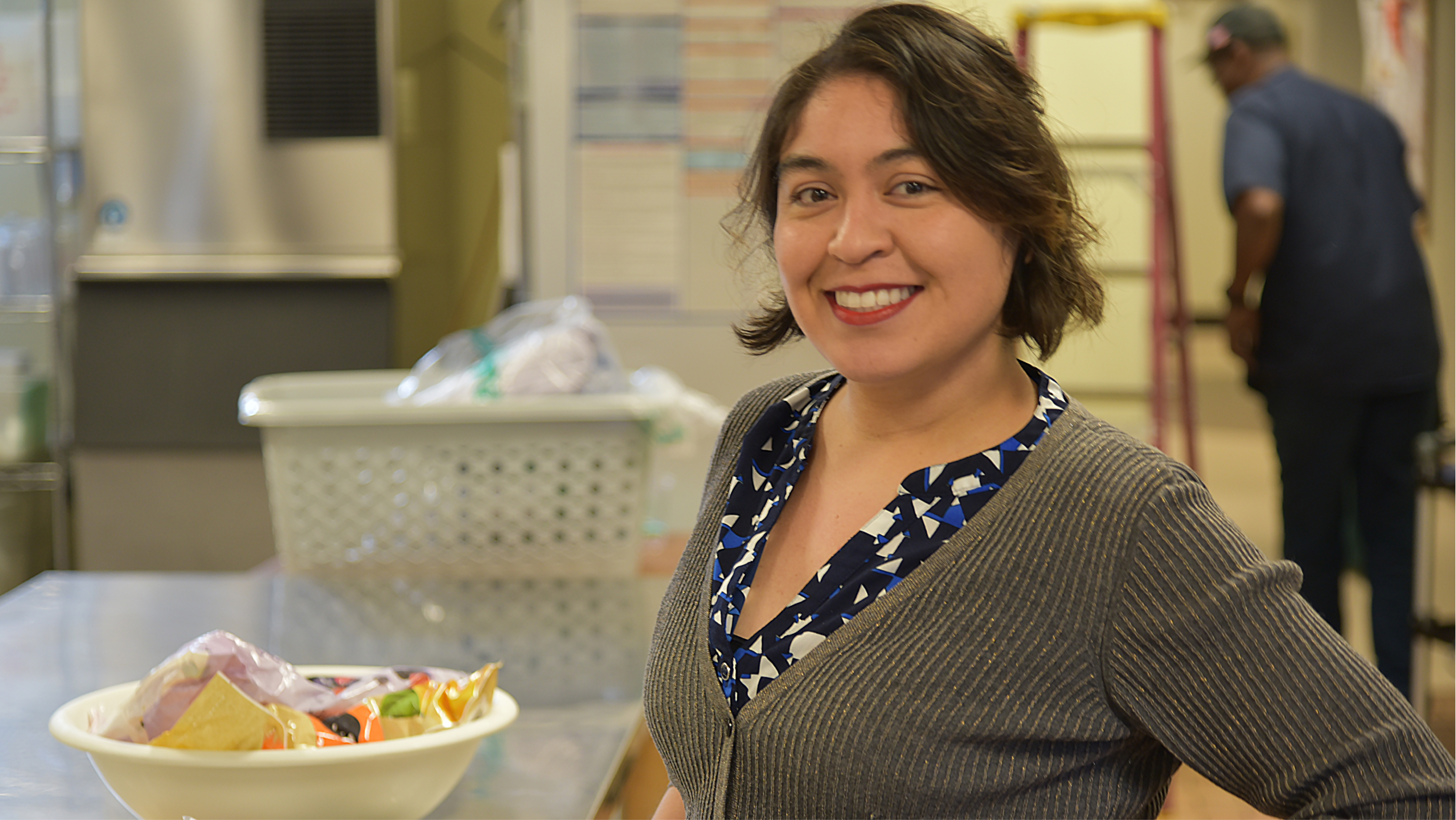 A smiling female staff member in a grey cardigan standing in a campus workspace.