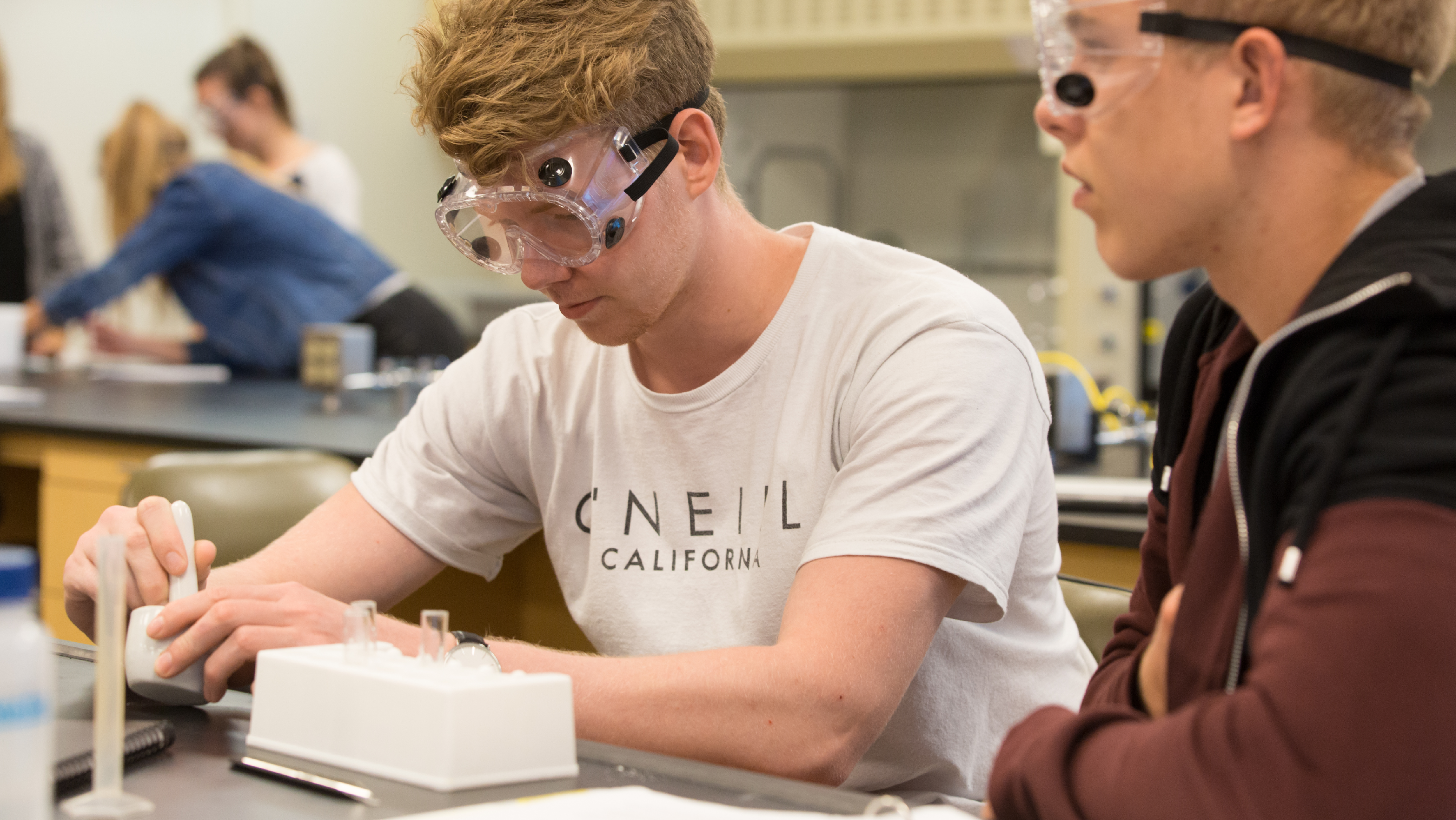 Four students wearing safety goggles closely examining a test tube containing a liquid solution.