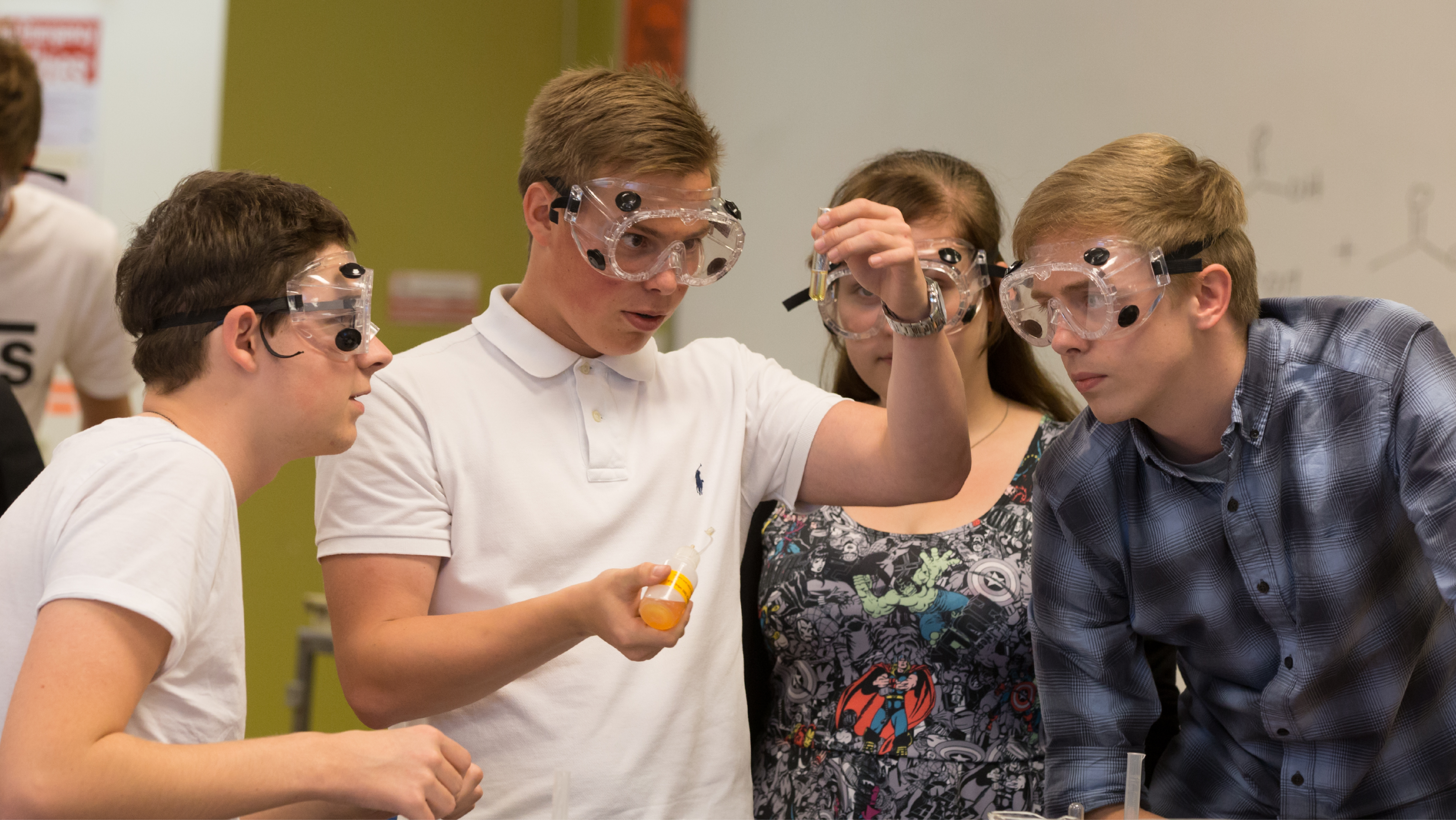 Three students in safety goggles observing a chemical solution in a test tube together.